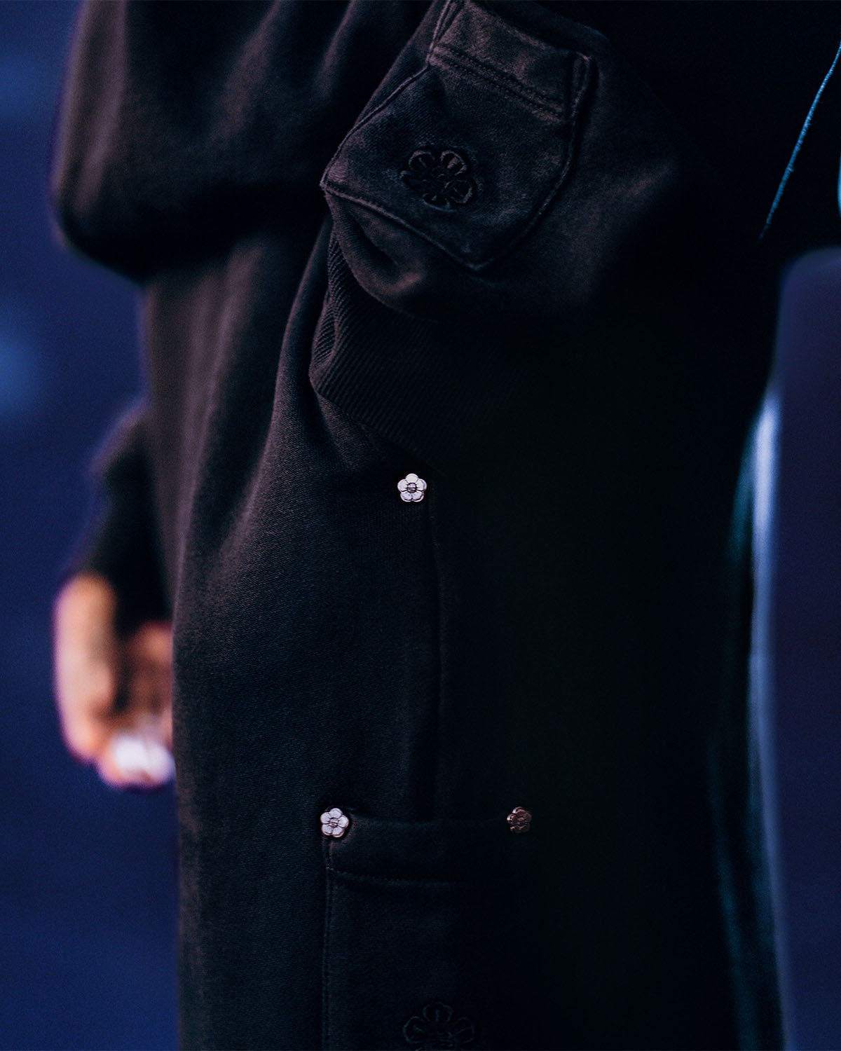 A close-up of a person wearing the Sundae School FADE BLACK WRAP SWEATBAJI, a hanbok-inspired dark long-sleeved piece with decorative buttons and embroidered floral details on the sleeve and pocket against a blurred background.