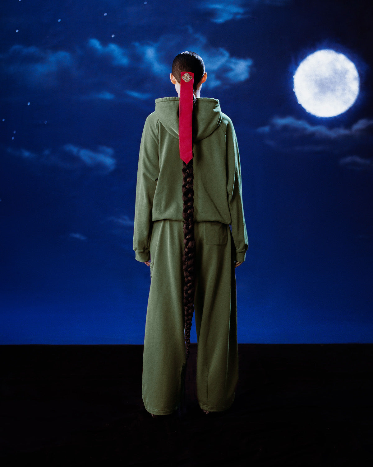 A person in the Sundae School KHAKI HANBOK HOODIE, featuring a signature stash pocket, stands with their back to the camera, long braid tied with a red ribbon, while a full moon glows in the cloudy night sky behind them.