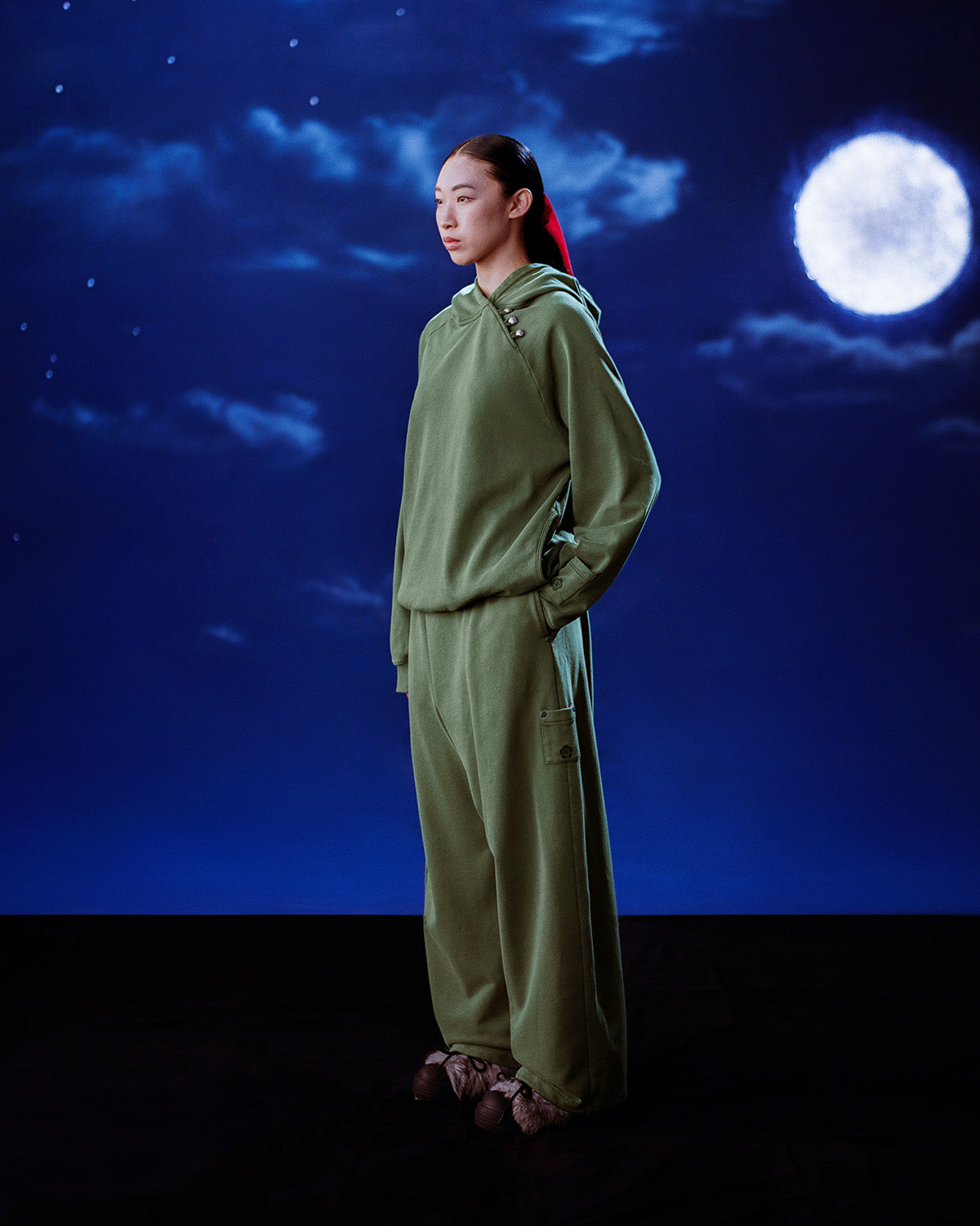 A person stands in profile wearing the loose-fitting Sundae School KHAKI HANBOK HOODIE with hands in pockets, set against a glowing night sky with clouds and a full moon over a dark, textured ground.