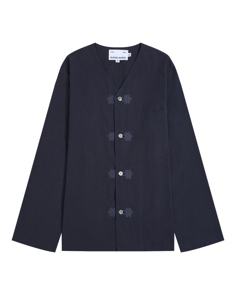 The MAEHWA COLLARLESS SHIRT by Sundae School is a navy blue, long-sleeve mid-layer featuring a V-neck, frog closures, and a single chest pocket. This versatile unisex piece is displayed laid flat on a white background.