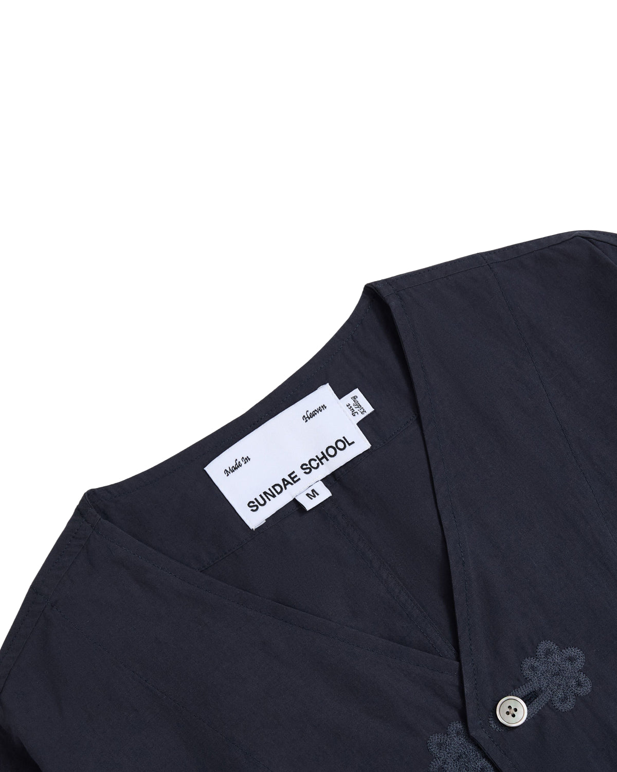 A close-up of the MAEHWA COLLARLESS SHIRT by Sundae School, a dark navy, collarless linen unisex mid-layer featuring delicate Maehwa floral embroidery near the neckline.