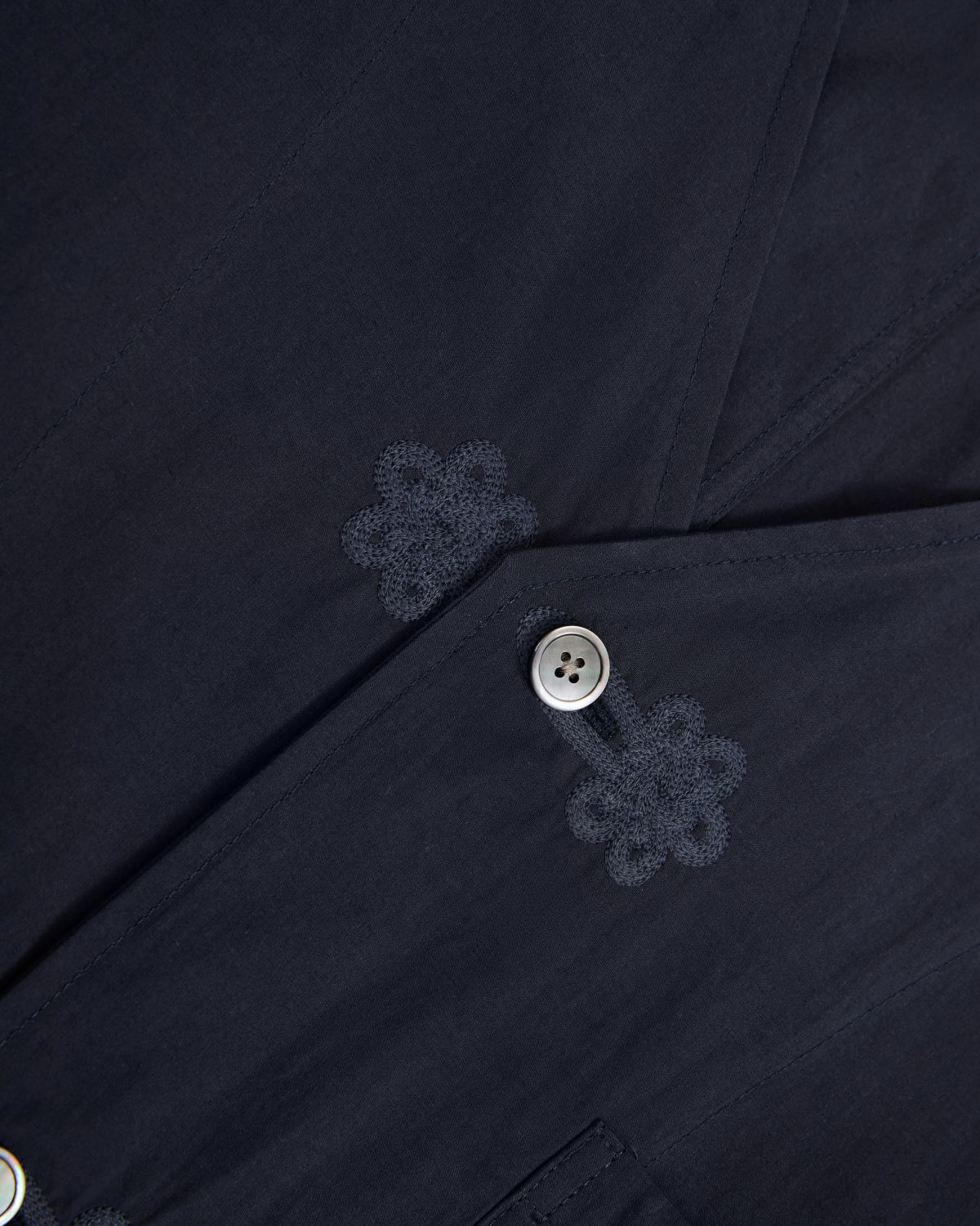 Close-up of the MAEHWA COLLARLESS SHIRT by Sundae School in dark navy, featuring two buttons and intricate Maehwa embroidery near the buttonholes. The smooth, finely detailed fabric enhances the refined aesthetic of this unisex mid-layer.