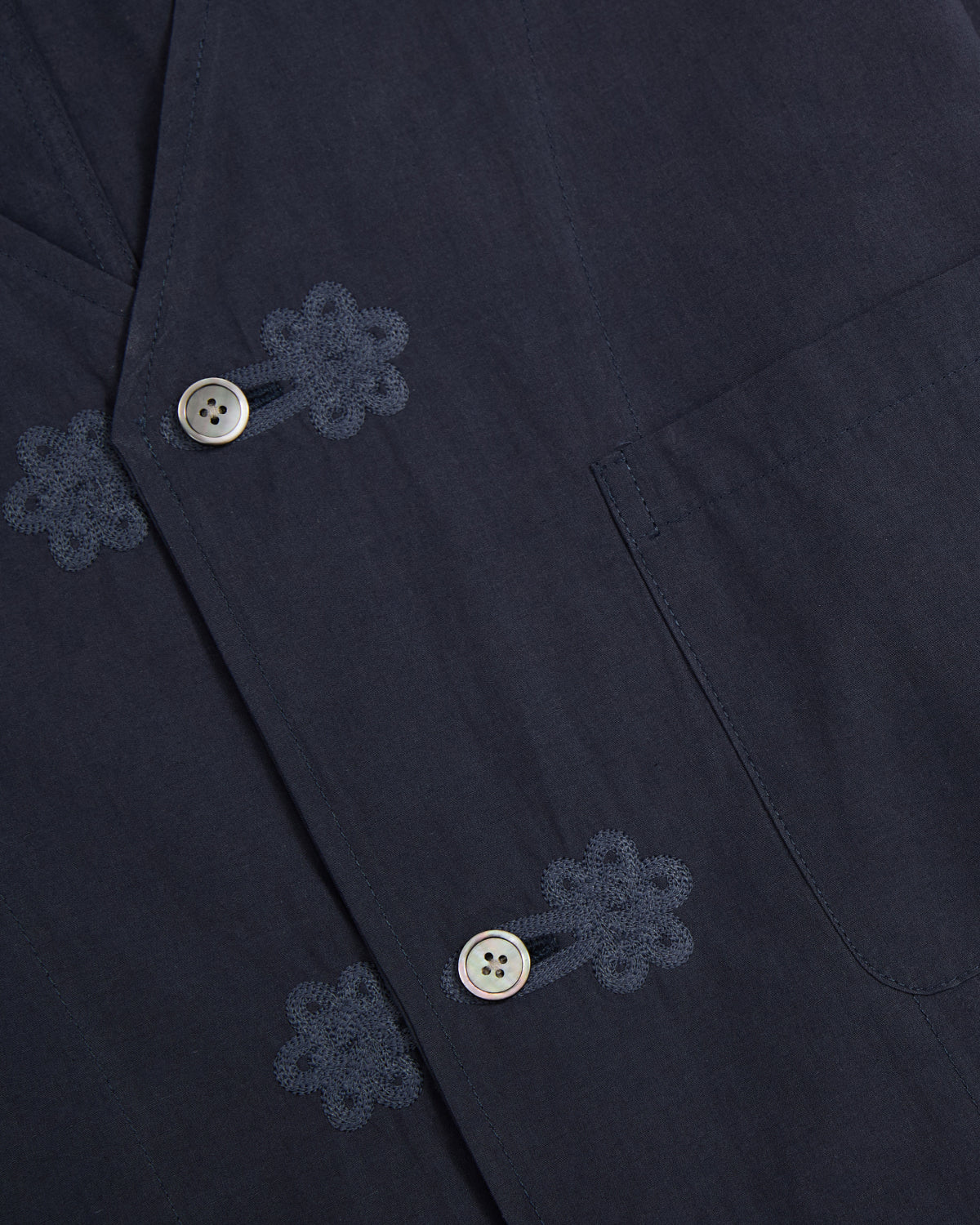 Close-up of the Sundae School MAEHWA COLLARLESS SHIRT in dark blue, a unisex mid-layer featuring a pocket, light-colored buttons, and decorative Maehwa embroidery near the buttonholes.