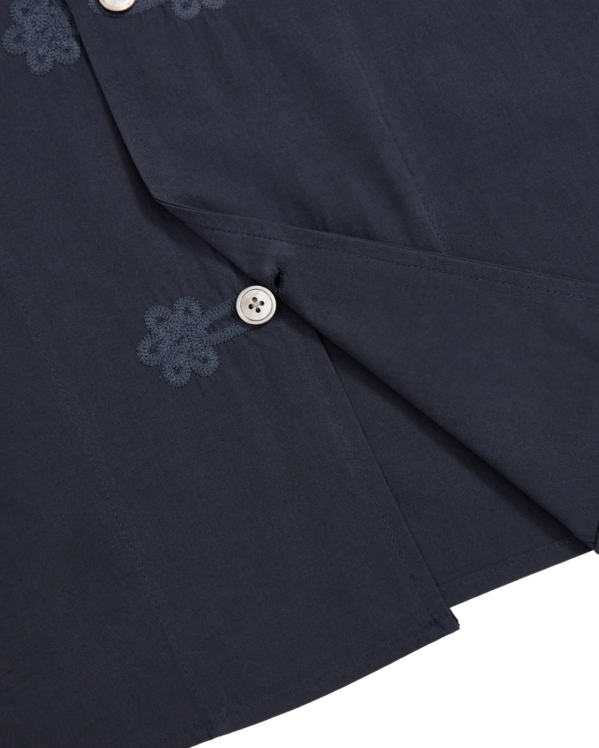 Close-up of the Sundae School MAEHWA COLLARLESS SHIRT, crafted in dark blue linen without a collar and accented by white buttons. Near the buttonholes, delicate Maehwa embroidery adds a unique touch. The slightly open weave reveals an inner layer, making this unisex shirt an ideal mid-layer for versatile styling.