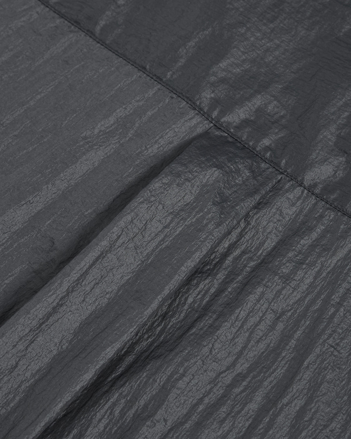 Close-up of the dark, crinkled fabric from Sundae School’s MAEHWA EMBROIDERED SHEER SHIRT, a unisex going-out piece, featuring a visible horizontal stitched seam and showcasing textured folds under soft lighting.