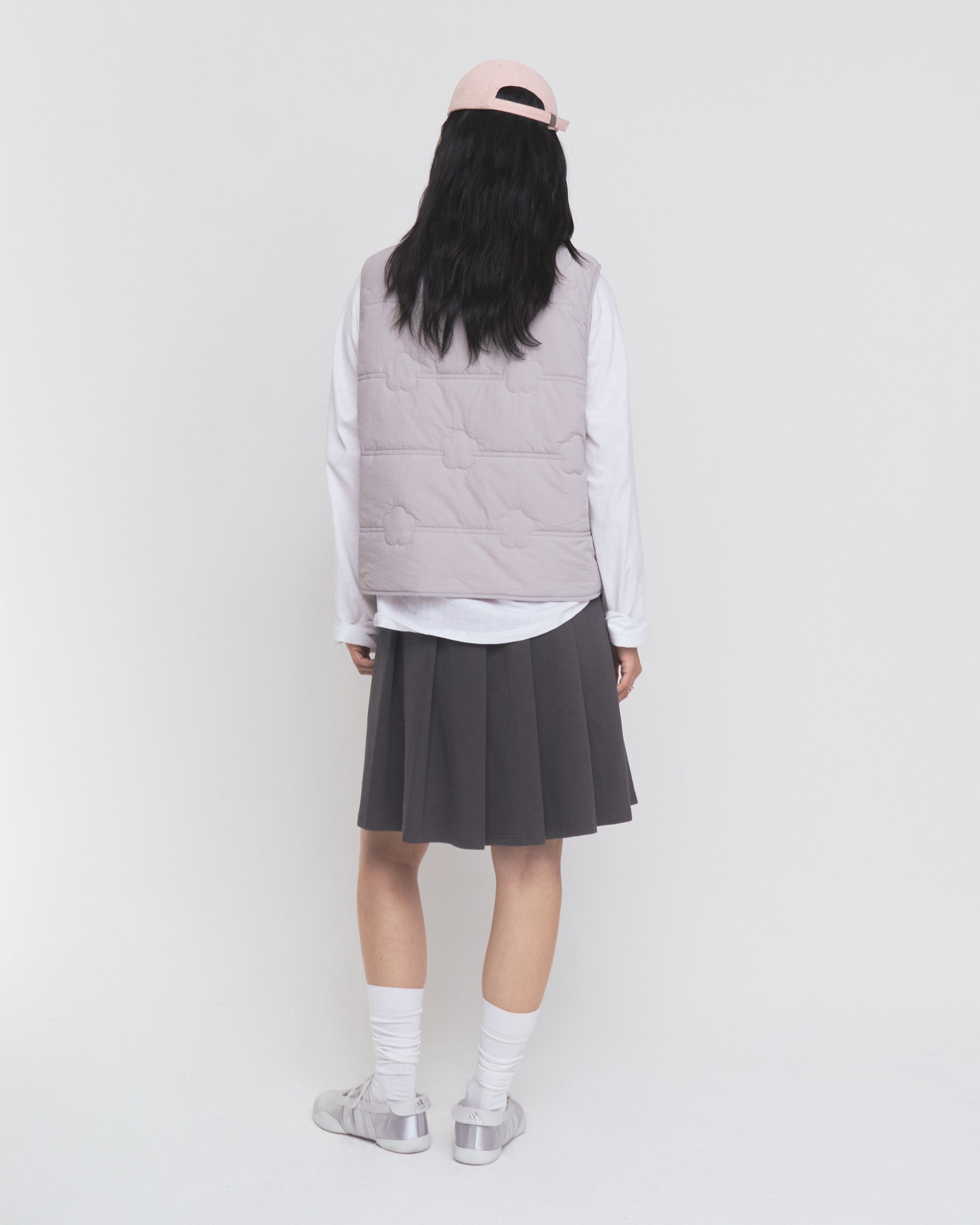 A person stands facing away against a plain white background, wearing a pale pink cap, the Sundae School BABY PINK MOMO REVERSIBLE FLEECE VEST, a white long-sleeve shirt, a dark pleated skirt, white socks, and silver sneakers.