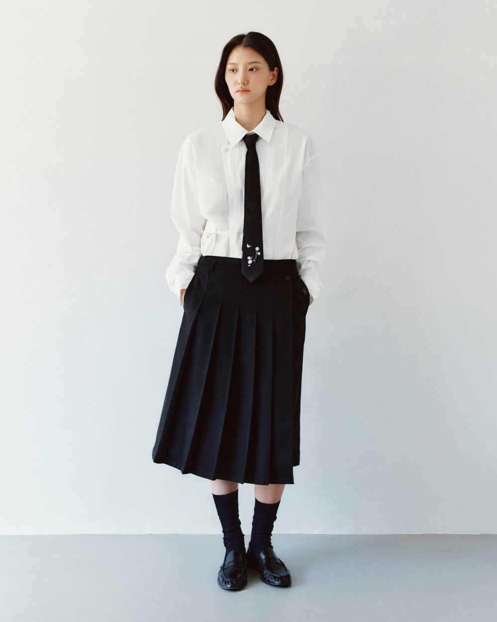 A person wears a white button-down shirt, a thin black necktie with floral embroidery, and a BLACK MOON WRAP MIDI SKIRT featuring an asymmetric pleated design.