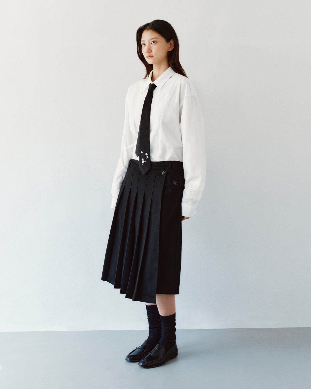 A person wears a white button-down shirt and a thin black necktie with a BLACK MOON WRAP MIDI SKIRT, an asymmetric pleated style featuring an adjustable waist belt.