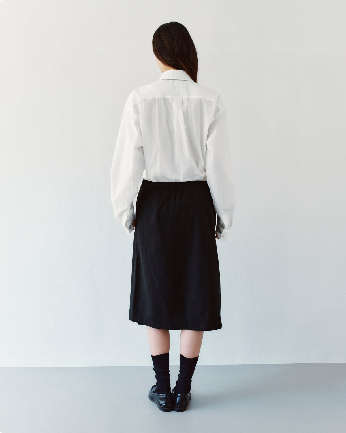 Back view of a person wearing a white collared shirt tucked into a black Sundae School BLACK MOON WRAP MIDI SKIRT, black socks, and dark loafers.