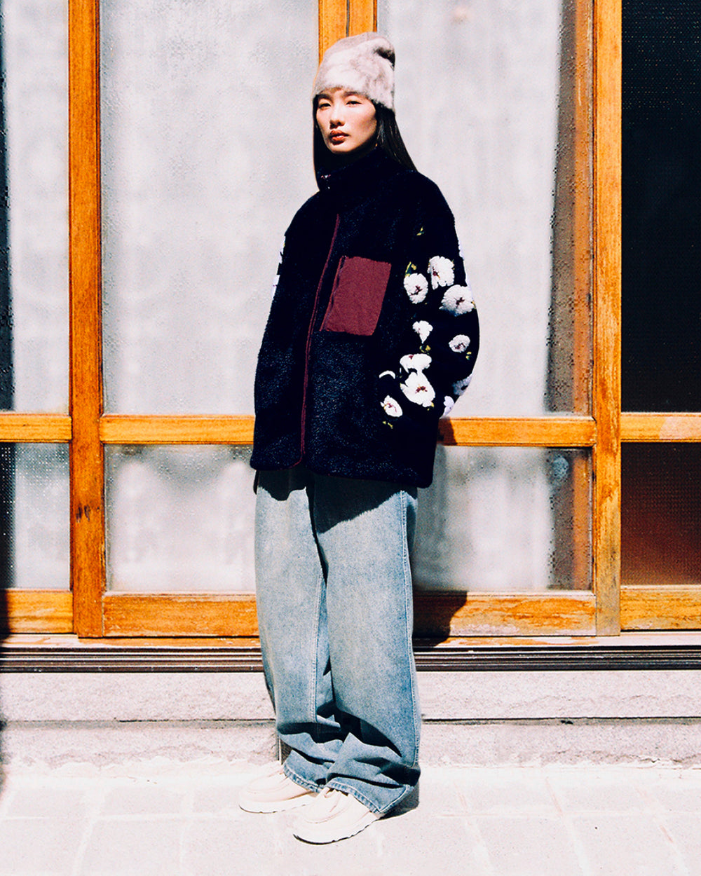 A woman stands outside in front of large wooden-framed windows, wearing the Sundae School MUGUNGHWA FLEECE—a sherpa fleece jacket featuring floral sleeves inspired by the Korean national flower—paired with loose jeans, white sneakers, and a light furry hat.