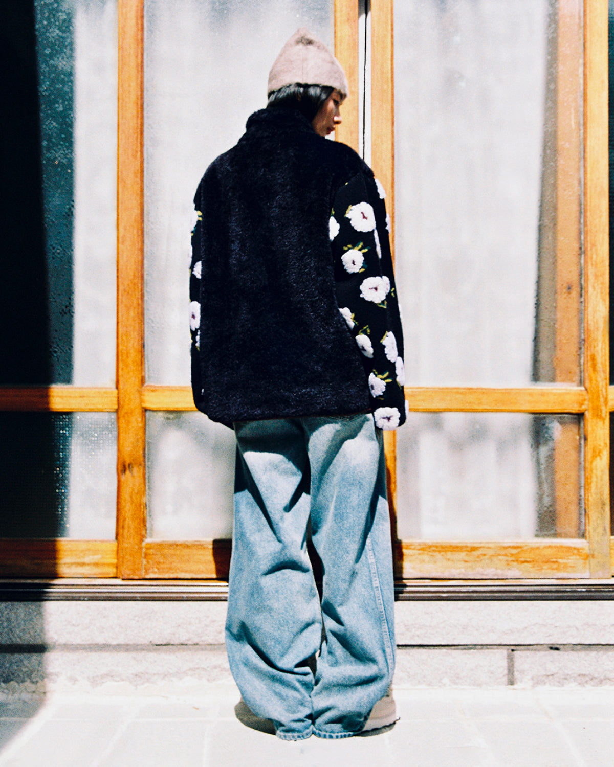 A person stands with their back to the camera, wearing a gray beanie and Sundae School’s MUGUNGHWA FLEECE—a black sherpa jacket featuring floral sleeves inspired by the Korean national flower—paired with loose-fitting blue jeans, in front of a wooden-framed glass window.