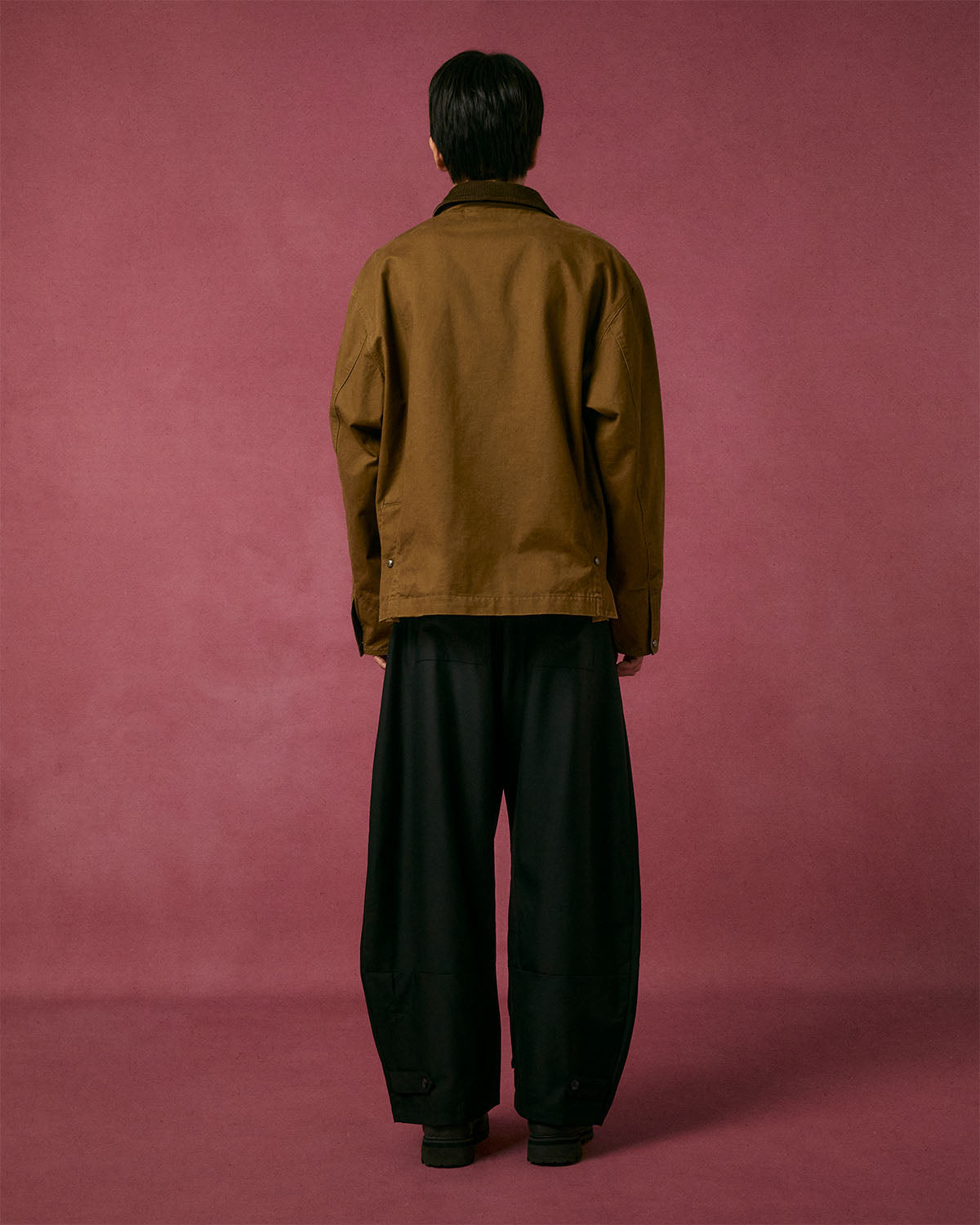A person with short dark hair faces a pinkish-red wall, wearing Sundae School's TAN KNOT BARN JACKET and loose black pants. Their back is to the camera, hands relaxed at their sides.
