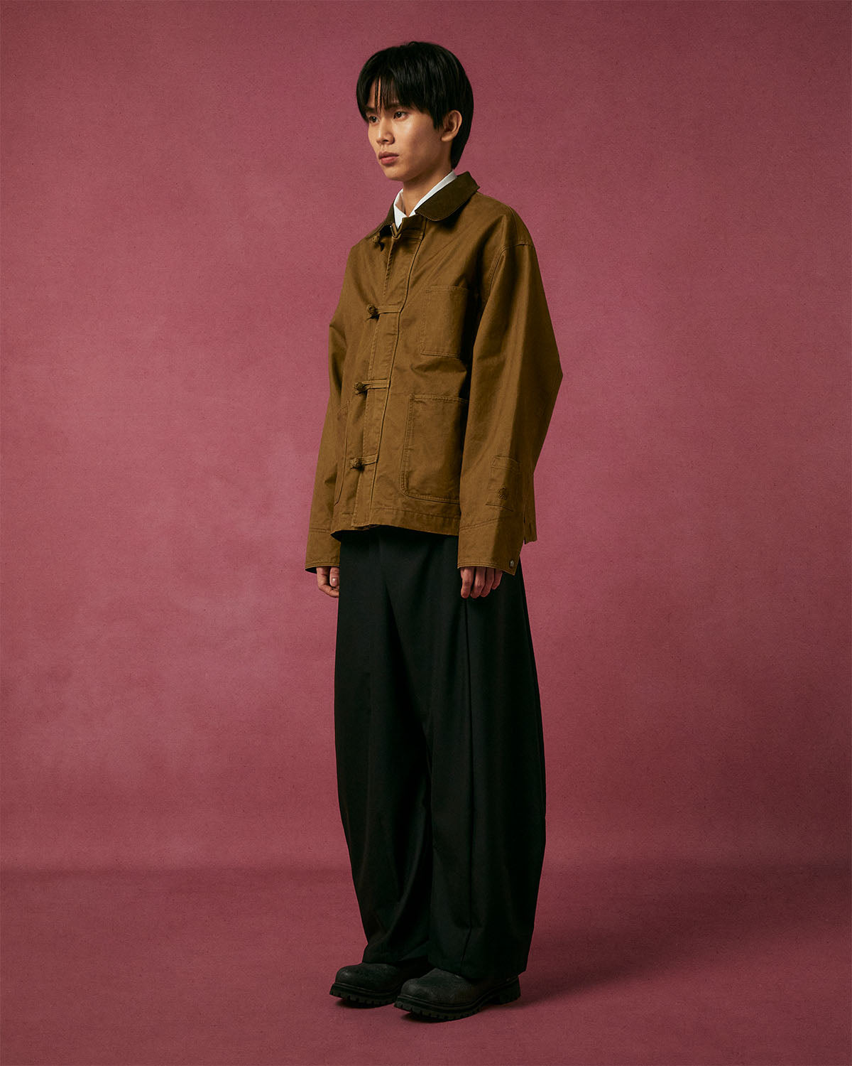 A person with short dark hair stands against a pink backdrop, wearing the Sundae School TAN KNOT BARN JACKET over a white shirt, wide black pants, and black shoes. They face slightly to the side with a neutral expression.