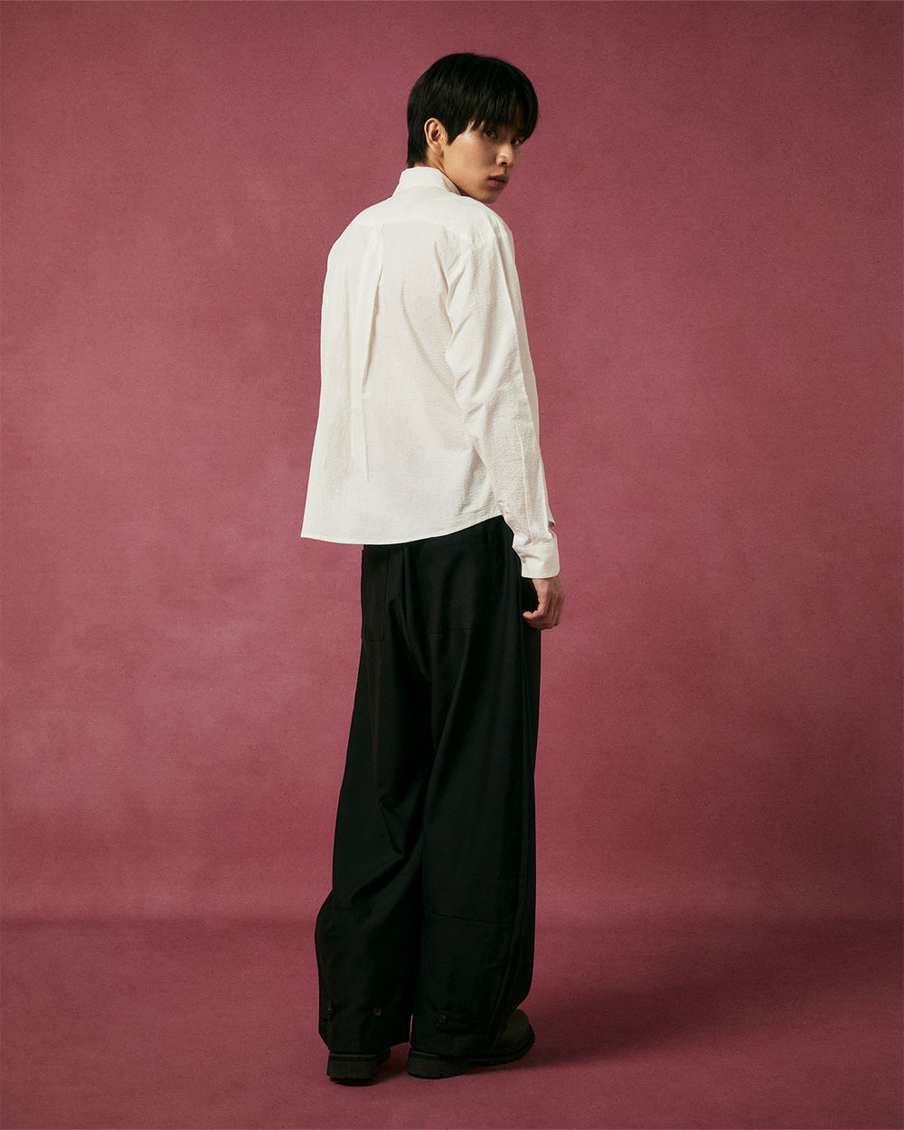 Against a maroon backdrop, a person faces away but glances back, wearing the IVORY TIE GORUM SHIRT by Sundae School with wide-leg black pants and dark shoes.