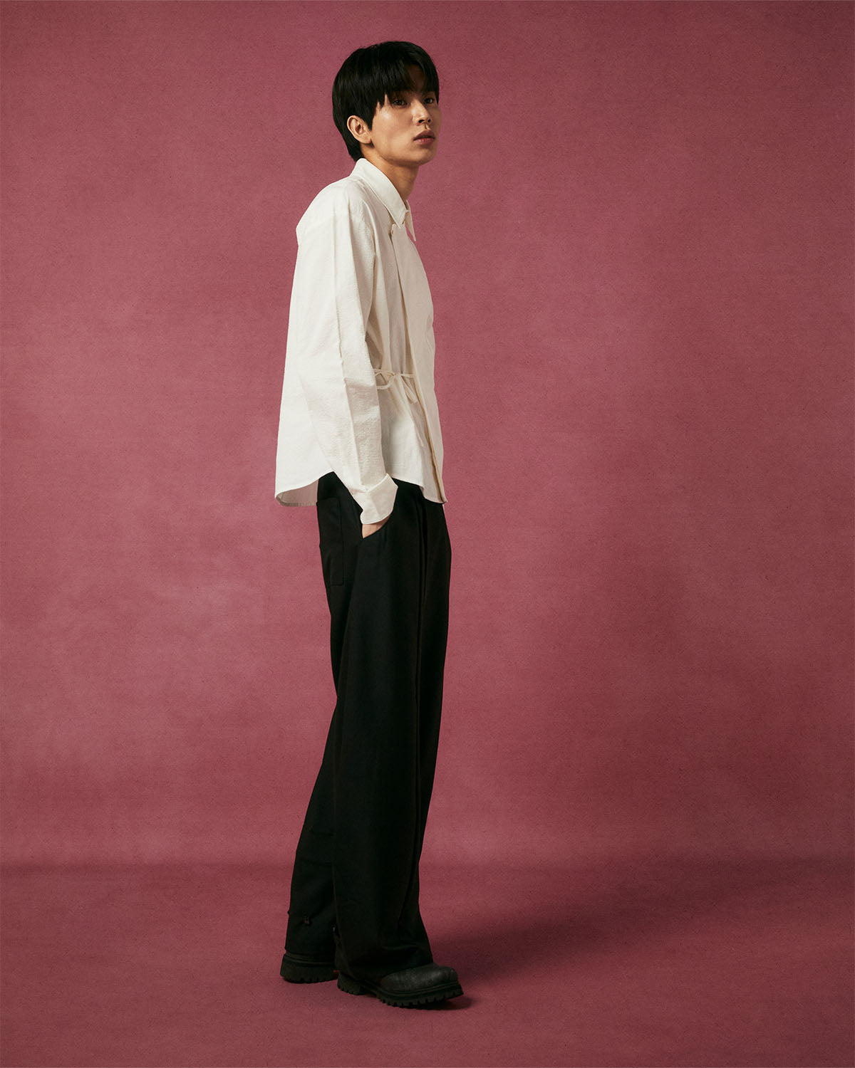 A person with short dark hair stands sideways on a maroon background, wearing Sundae School's IVORY TIE GORUM SHIRT, along with black pants and shoes, hands in pockets, looking slightly upward.