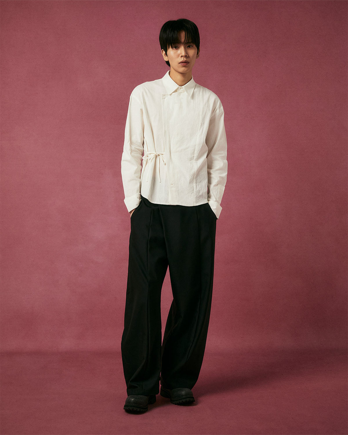 A person stands against a mauve background wearing Sundae School’s IVORY TIE GORUM SHIRT, styled with loose black pants and black shoes. They have straight dark hair and a neutral expression.
