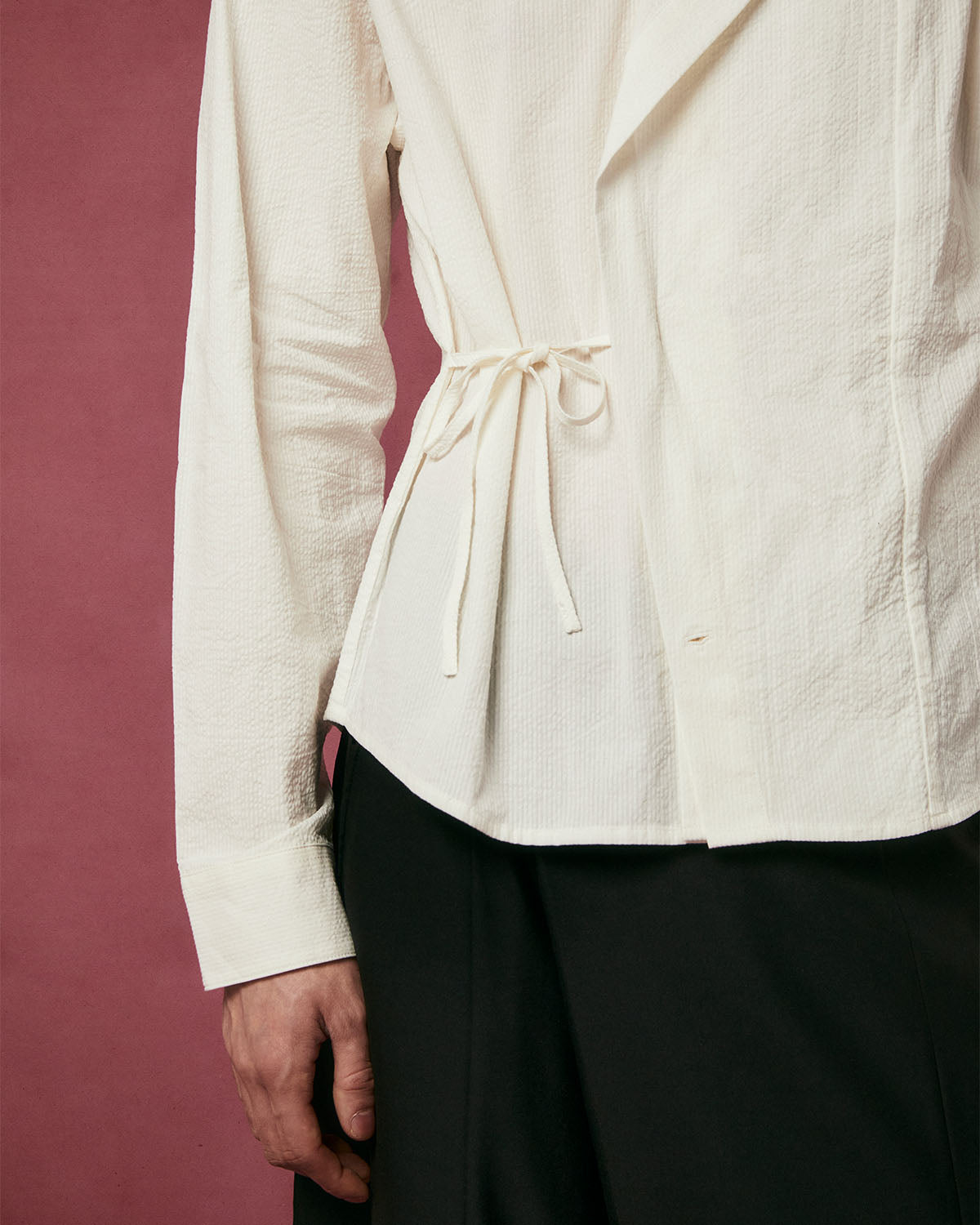 A person wears the Sundae School IVORY TIE GORUM SHIRT, featuring a back waist drawstring, paired with black pants against a dark pink background.