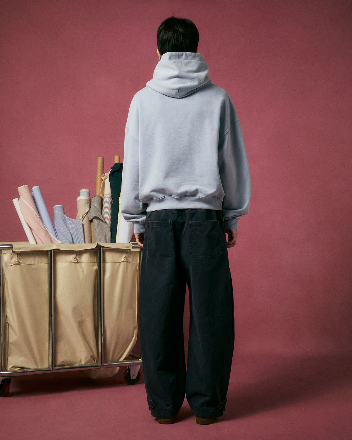 A person stands with their back to the camera, wearing the Sundae School SKY BLUE COCOON HOODIE with its oversized hood and loose dark pants, next to a large bin of rolled fabrics against a pink background.