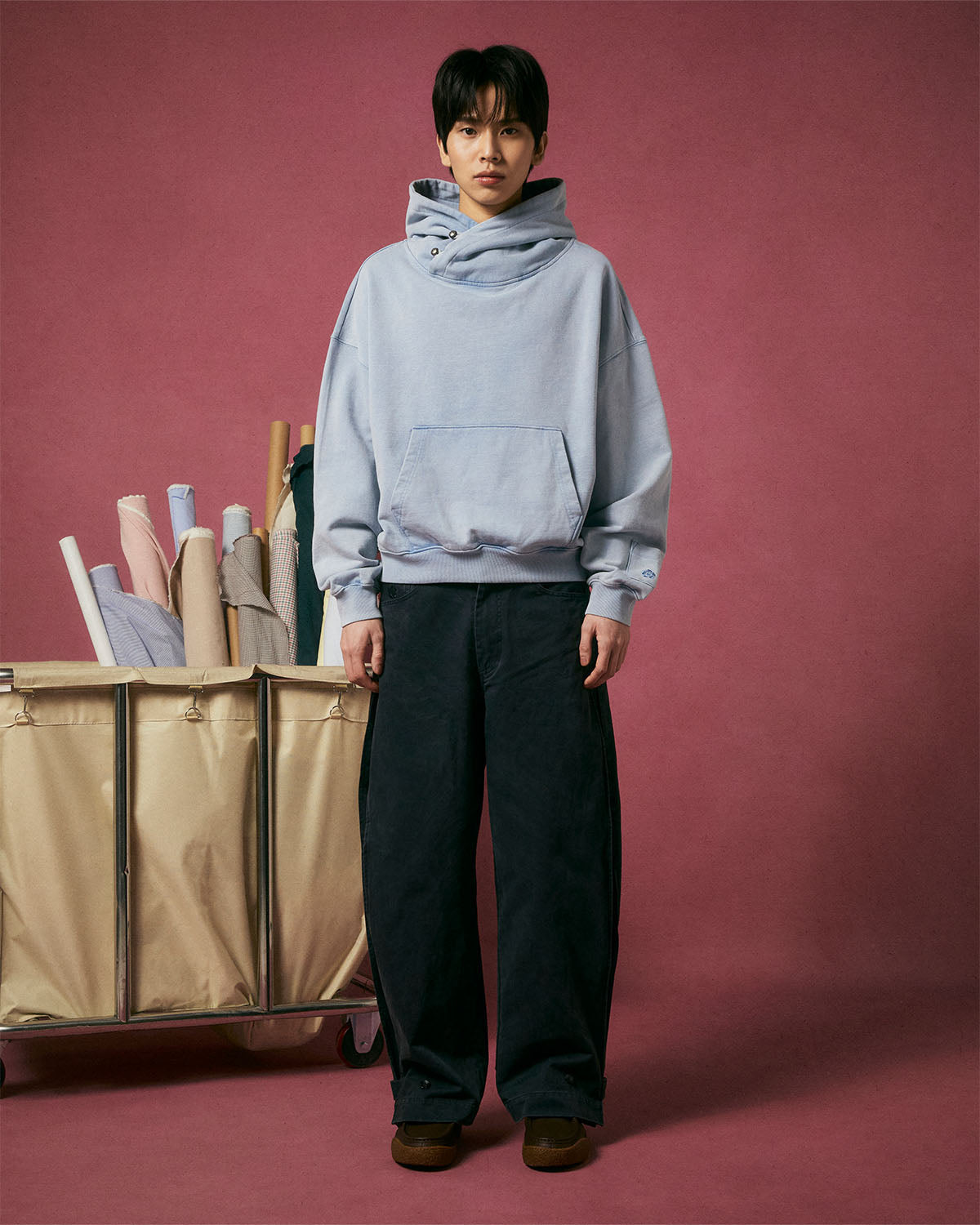 A person stands before a pink backdrop wearing the SKY BLUE COCOON HOODIE by Sundae School, paired with wide-leg black pants and brown shoes. Next to them, a beige cart displays colorful rolls of fabric.