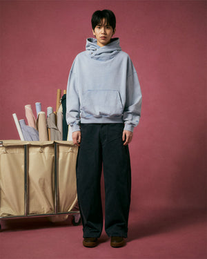 A person stands before a pink backdrop wearing the SKY BLUE COCOON HOODIE by Sundae School, paired with wide-leg black pants and brown shoes. Next to them, a beige cart displays colorful rolls of fabric.