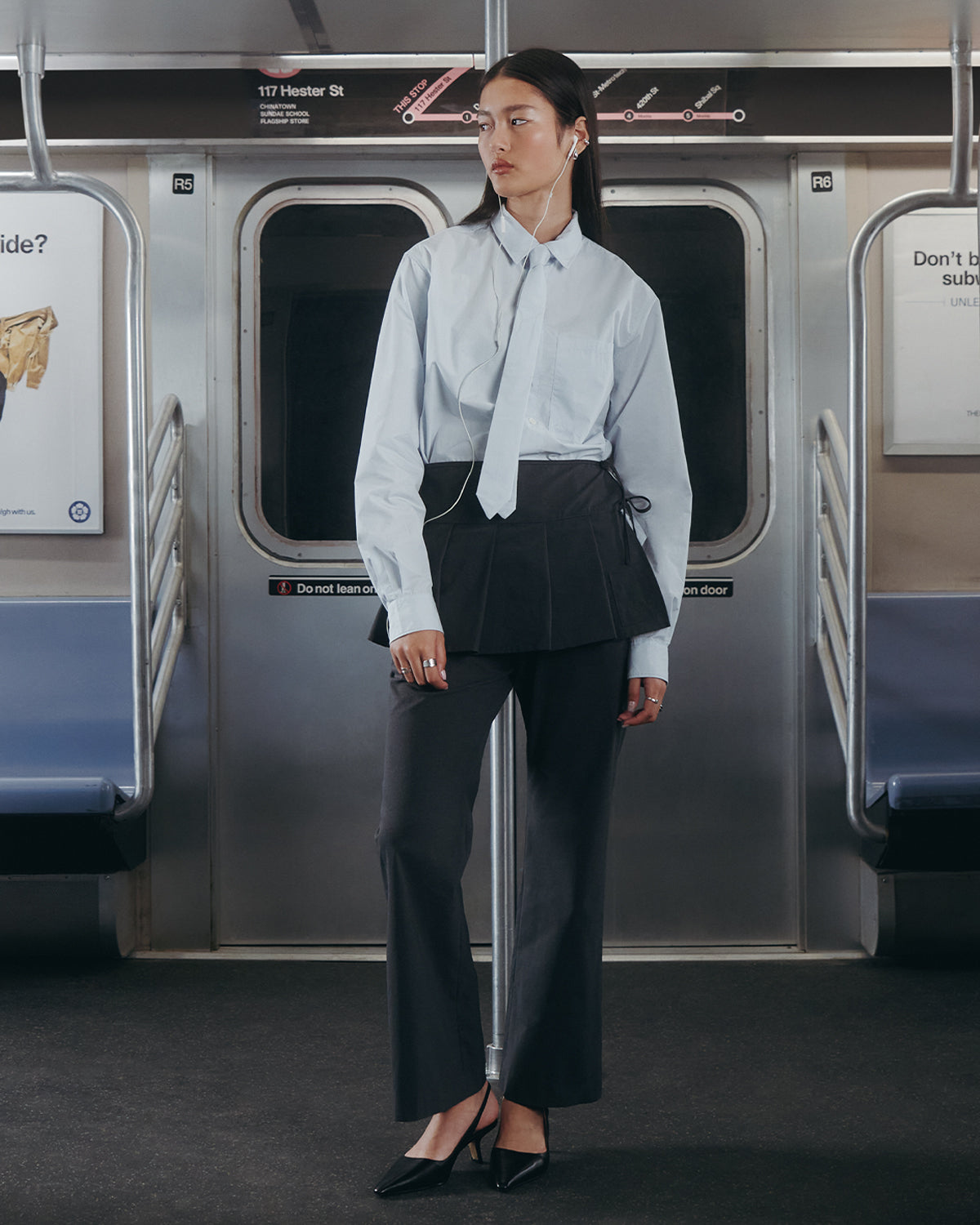 A woman in a Sundae School NECKTIE SHIRT with a matching removable tie, black skirt over dress pants, black heels, and earphones stands in a subway car, looking to the side with seats and ads visible behind her.