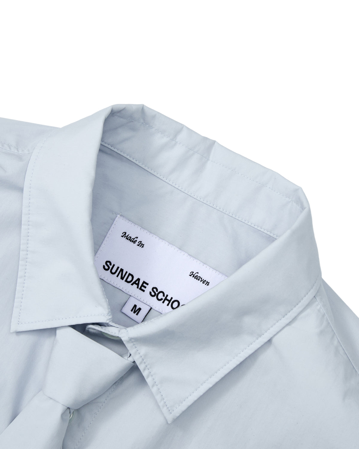 A NECKTIE SHIRT by Sundae School in light blue poplin, featuring a relaxed fit and a removable matching tie, is arranged neatly on a white background. The inside label reads “SUNDAE SCHOOL” and “Made In Heaven,” with the size marked as M.