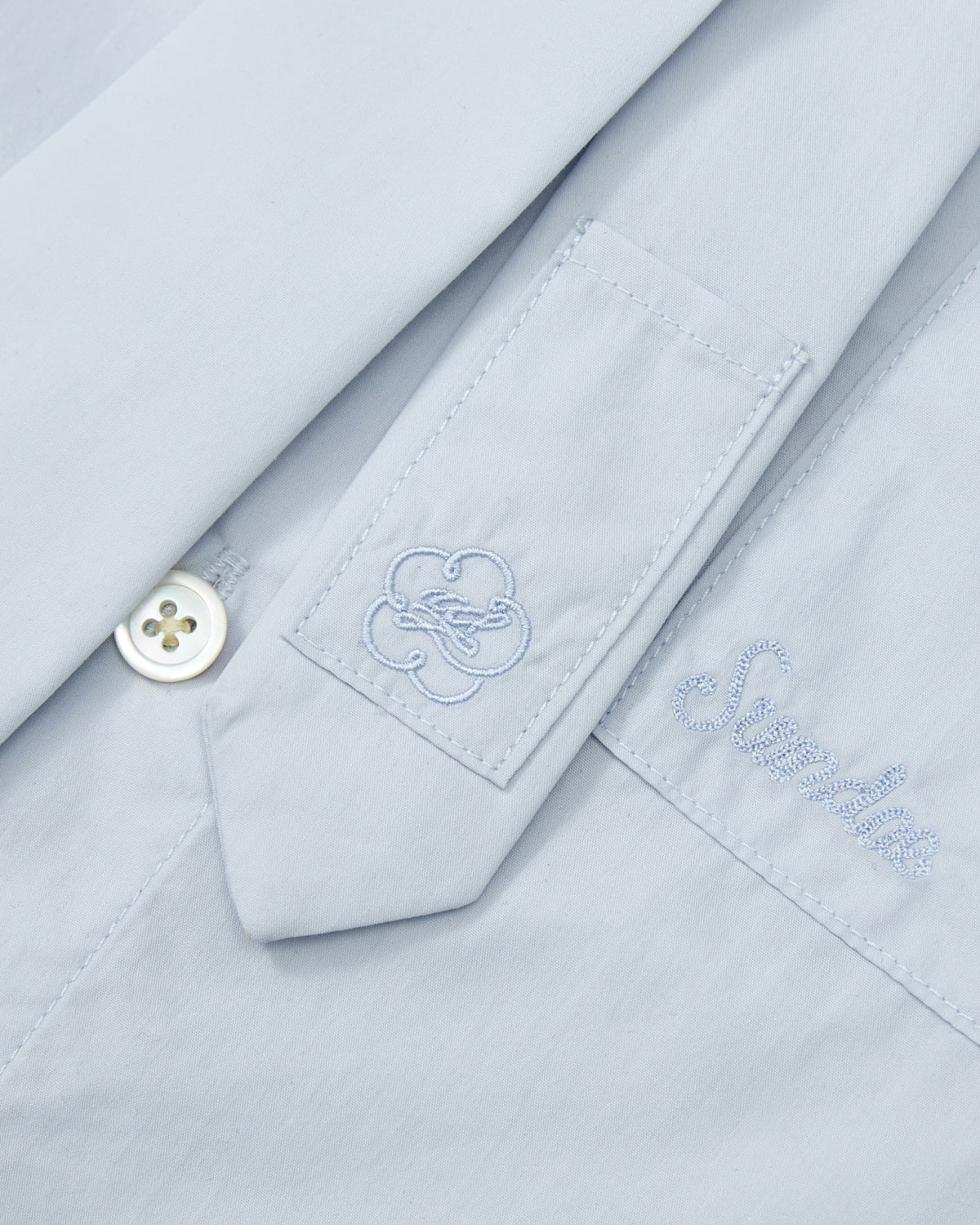 Close-up of the NECKTIE SHIRT by Sundae School in light blue, with a white button, embroidered swirl tab, 