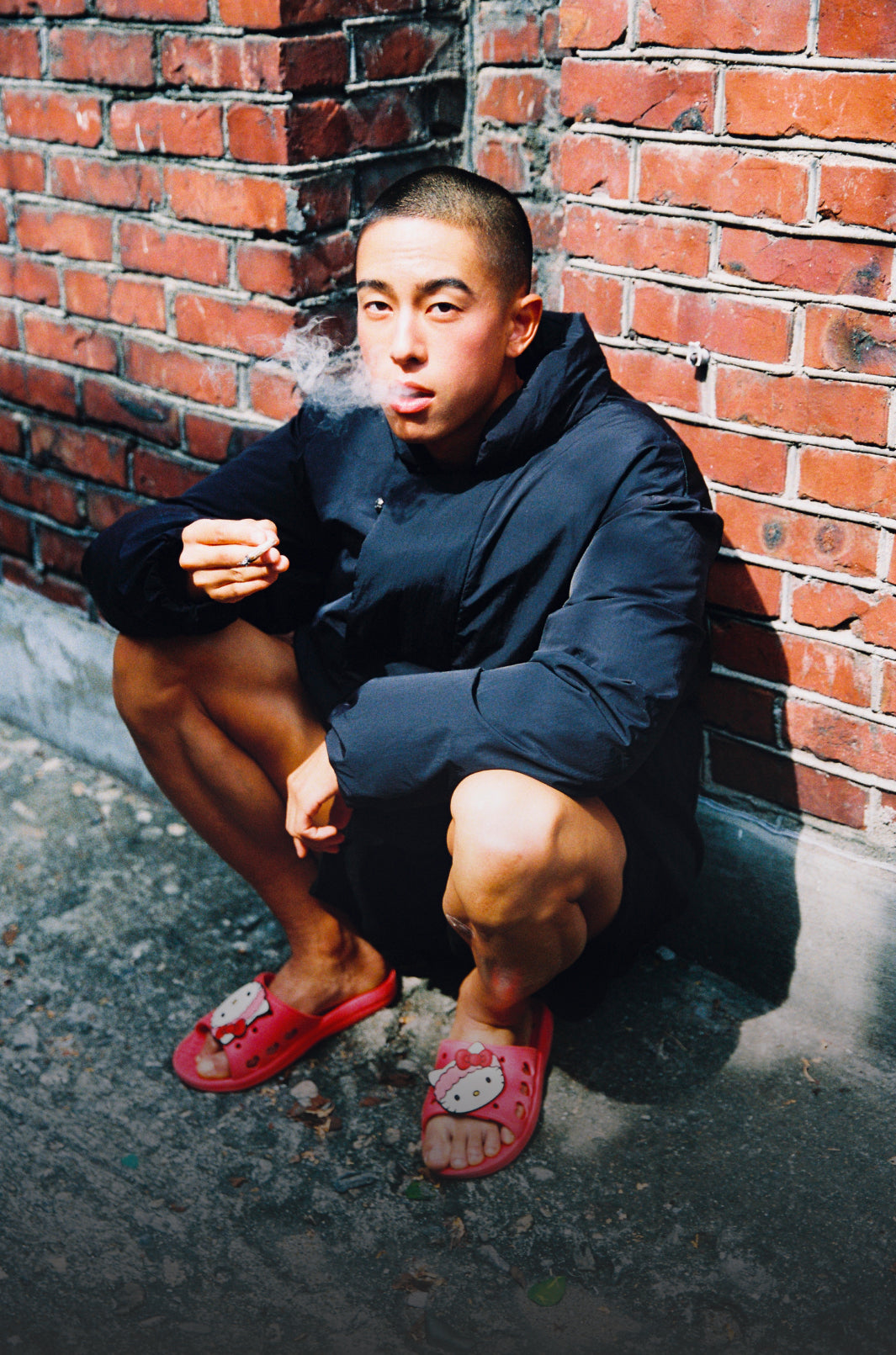A vertical image of a young man with a buzz cut squatting in front of a red brick wall on a sunny day. He is exhaling smoke and holding a lit cigarette in his right hand. He is wearing a black jacket, dark shorts, and red Hello Kitty slide sandals.