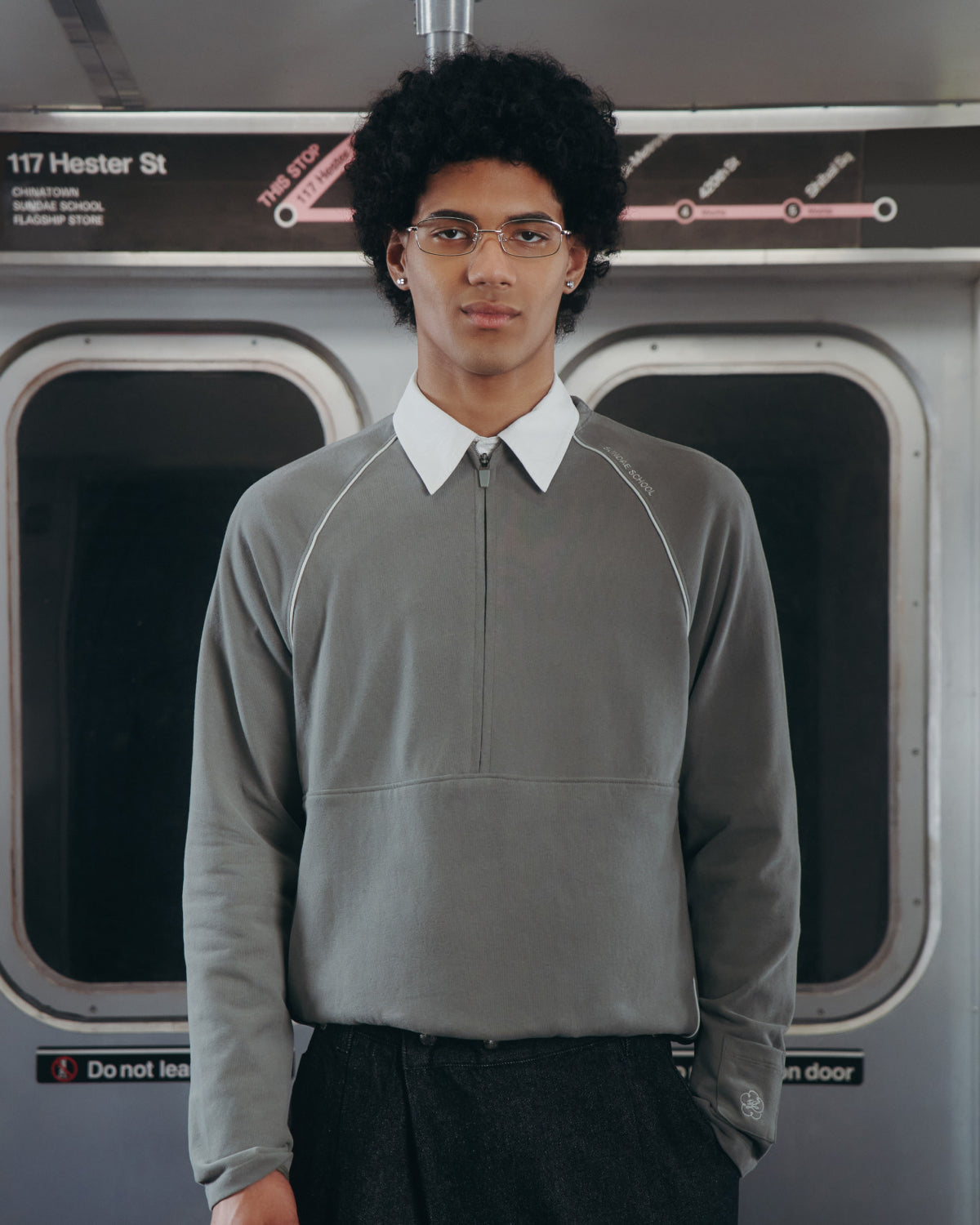 A person with short curly hair and glasses stands in a subway car with closed doors, wearing Sundae School’s KHAKI OFFICE QUARTER ZIP over a white collared shirt. The sign above reads 117 Hester St.