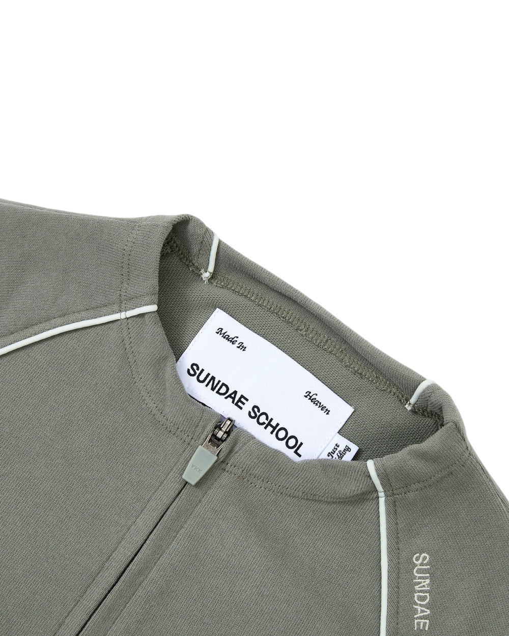 Close-up of the KHAKI OFFICE QUARTER ZIP by Sundae School, crafted from khaki-green cotton jersey with white piping detail. The inner label displays Sundae School and the text “Made In Heaven.” “SUNDAE” is printed near the collar, and a discreet stash pocket enhances the functionality of this quarter-zip design.