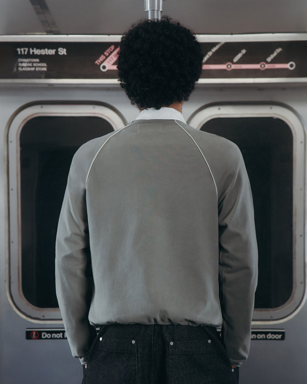 A person with curly hair, seen from behind in a Sundae School KHAKI OFFICE QUARTER ZIP, stands inside a subway train facing closed doors. Above the doors are a subway map and a sign that reads 117 Hester St.