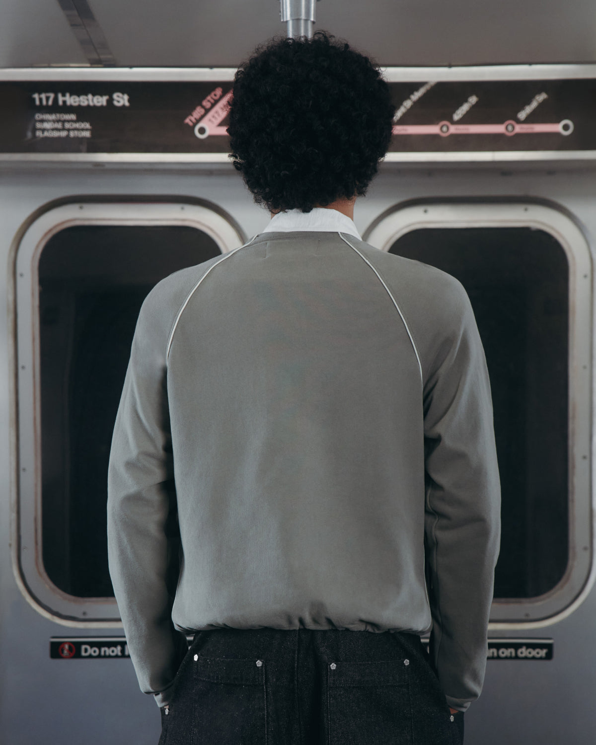 A person with curly hair, seen from behind in a Sundae School KHAKI OFFICE QUARTER ZIP, stands inside a subway train facing closed doors. Above the doors are a subway map and a sign that reads 117 Hester St.