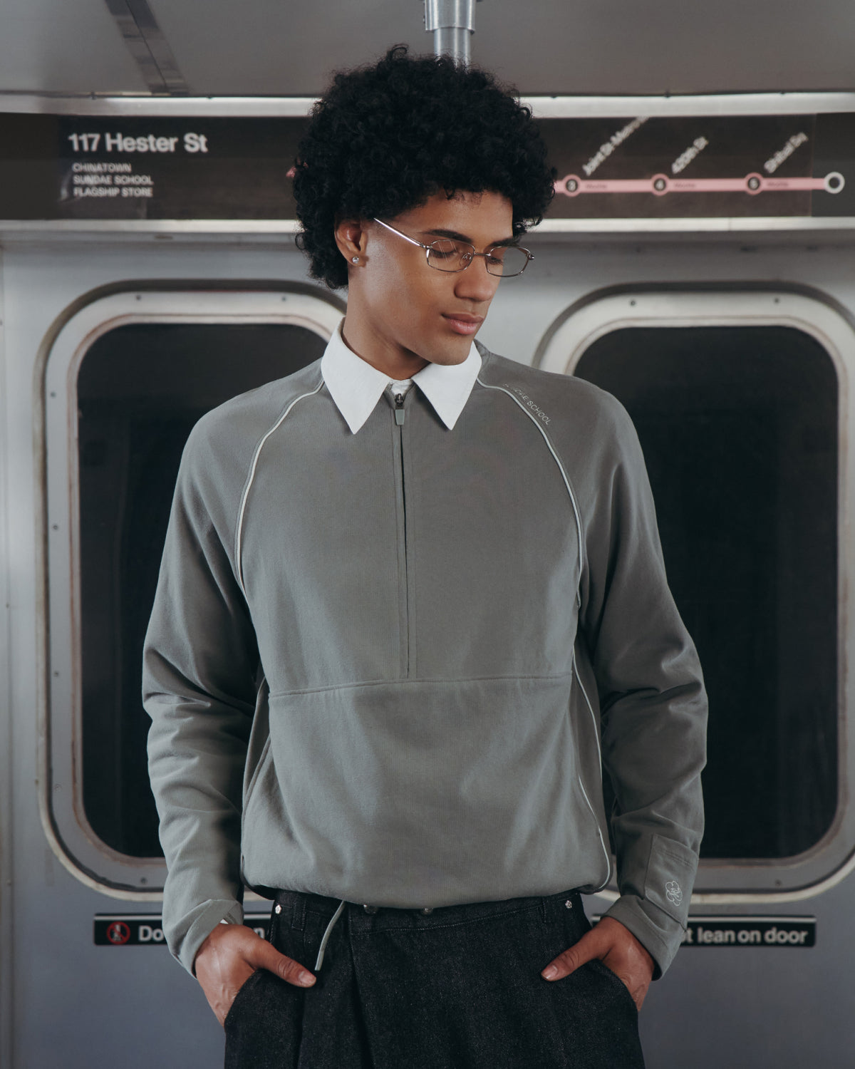 A person with curly hair and glasses wears a Sundae School KHAKI OFFICE QUARTER ZIP in a subway car, hands in pockets, with a subway map and "117 Hester St" sign visible behind them.
