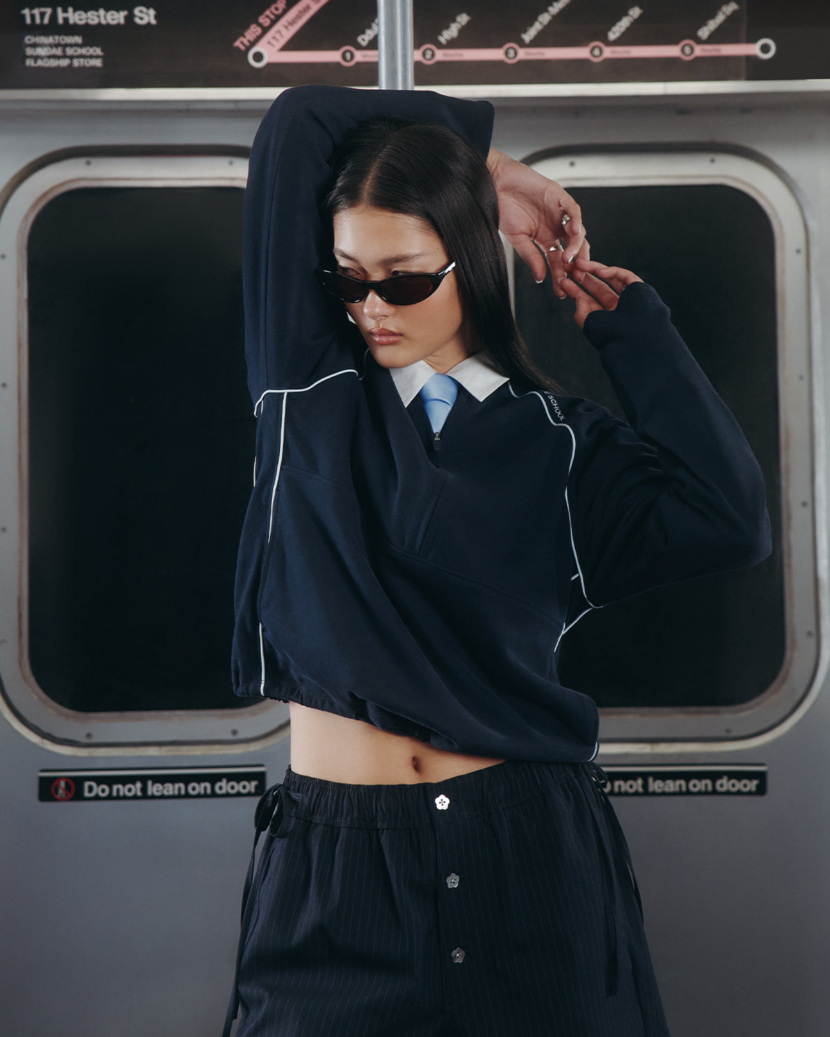 A person wearing the Sundae School NAVY OFFICE QUARTER ZIP and sunglasses poses with one arm raised inside a subway train, standing in front of closed doors with a 