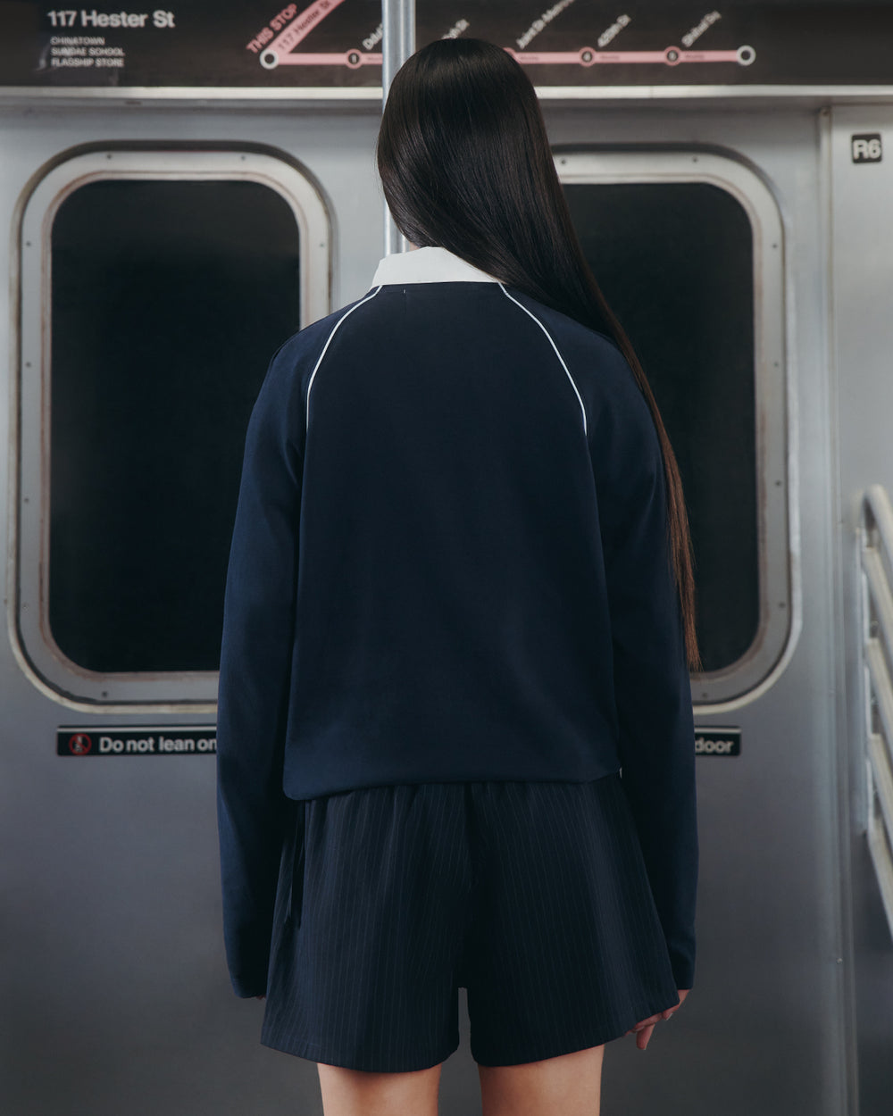 A person with long dark hair stands facing away in a subway car, wearing the Sundae School NAVY OFFICE QUARTER ZIP and shorts. Subway doors and signs for 117 Hester St and the R6 train are visible behind them.