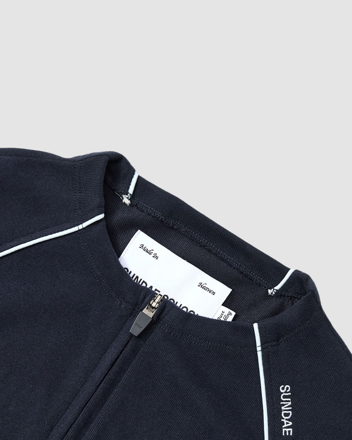 Close-up of the Sundae School NAVY OFFICE QUARTER ZIP, a navy blue cotton jersey with white piping and SUNDAE text on the collar. A white label appears inside the neck, shown against a light grey background.