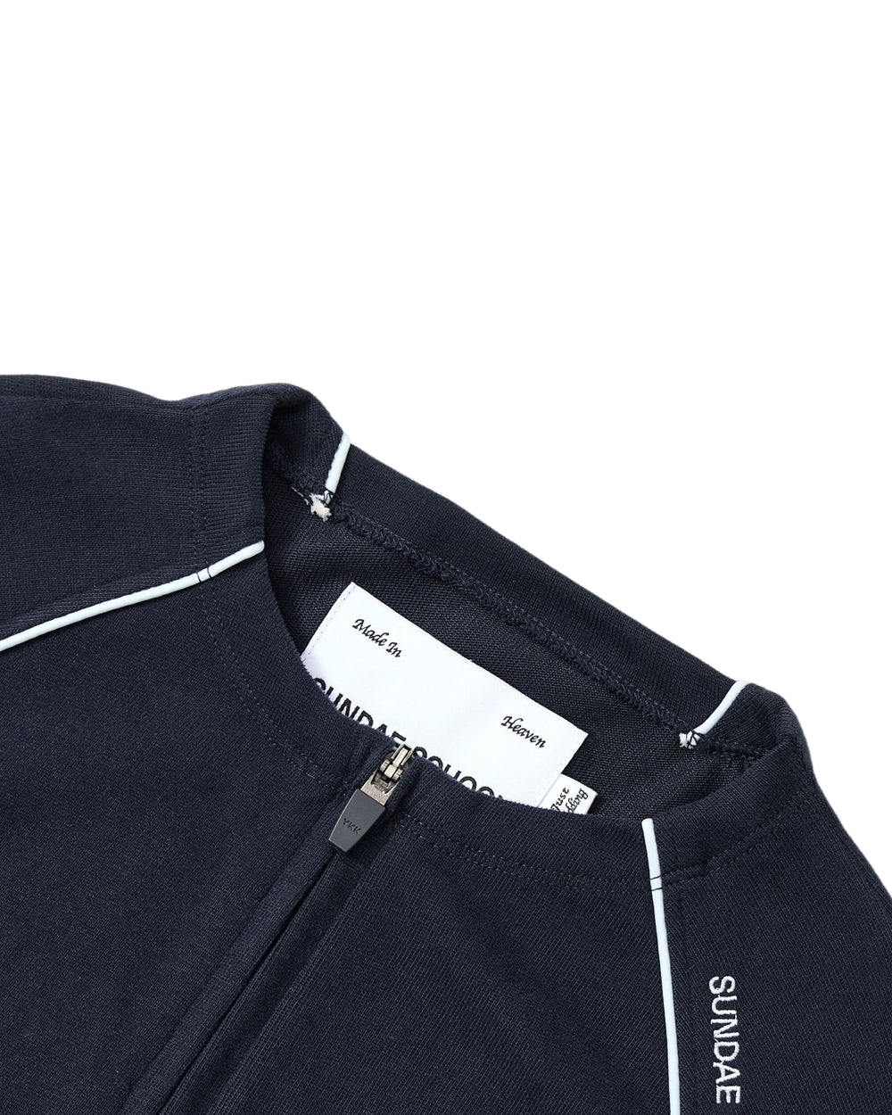 Close-up of the Sundae School NAVY OFFICE QUARTER ZIP, a navy blue cotton jersey zip-up featuring white piping and “SUNDAE” text on the collar. A white clothing label is visible inside near the zipper.