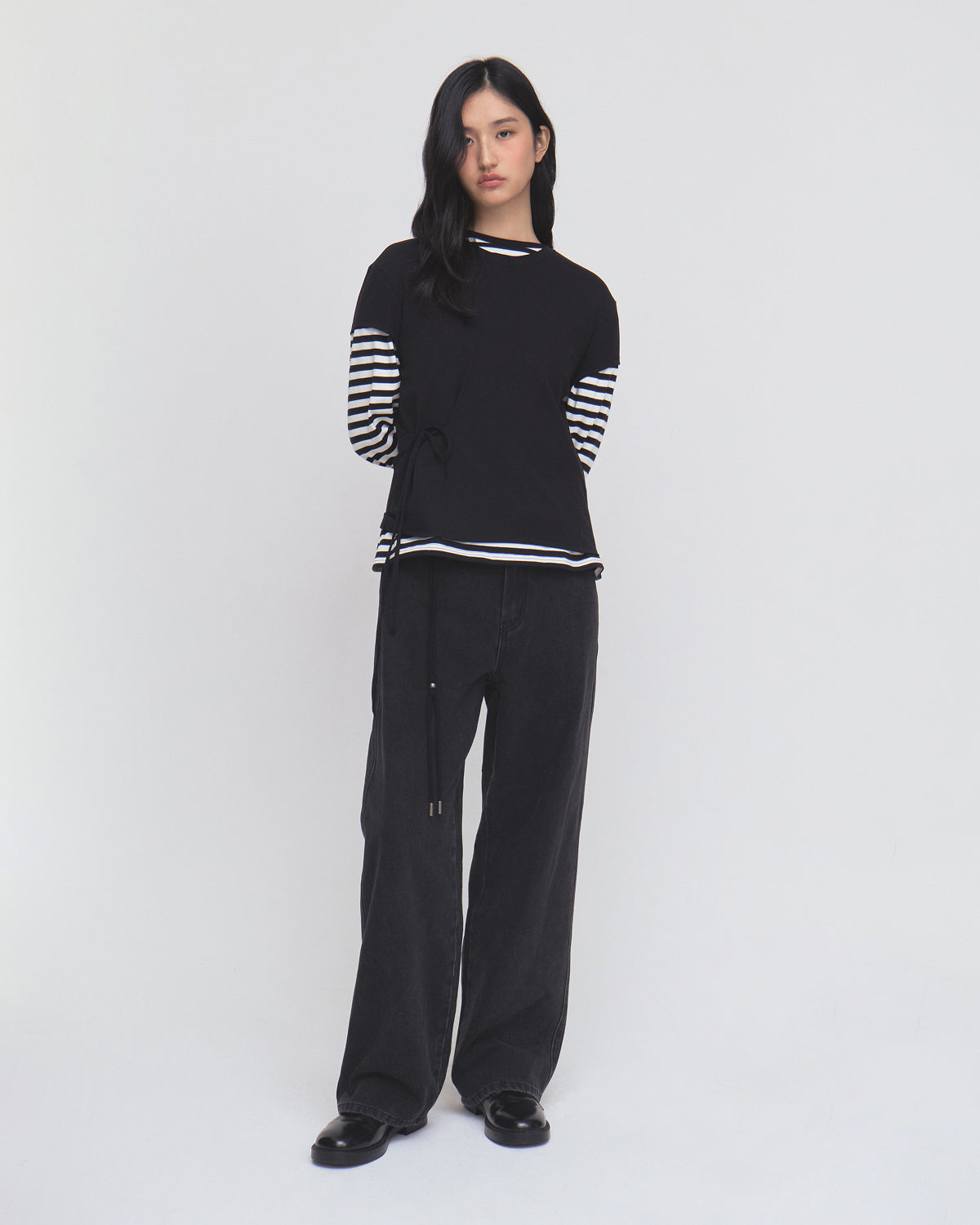 A woman stands against a plain white background, wearing the BLACK ORIGAMI GORUM TEE by Sundae School, which features black-and-white striped sleeves. She pairs the top with loose black pants and black shoes. Her long dark hair falls loosely, and she has a neutral expression.