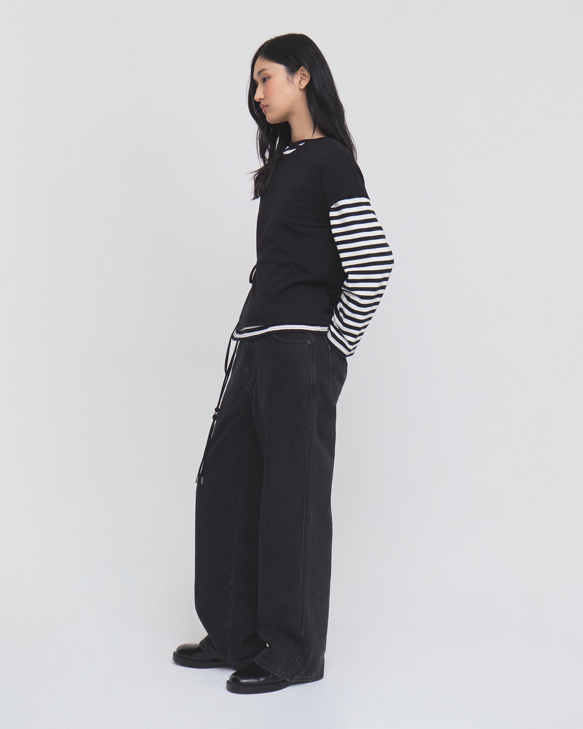 A woman stands sideways against a plain light background, wearing Sundae School’s BLACK ORIGAMI GORUM TEE—a black top with black-and-white striped long sleeves—paired with wide-leg black pants and black shoes.