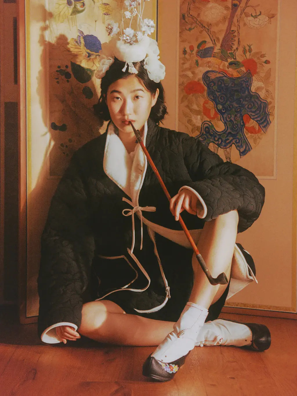 A person wearing a Sundae School Reversible Flower Quilted Fleece  and a decorative white floral headpiece sits on a wooden floor indoors, holding a long brown object. The background shows warm-toned wall panels with painted motifs.