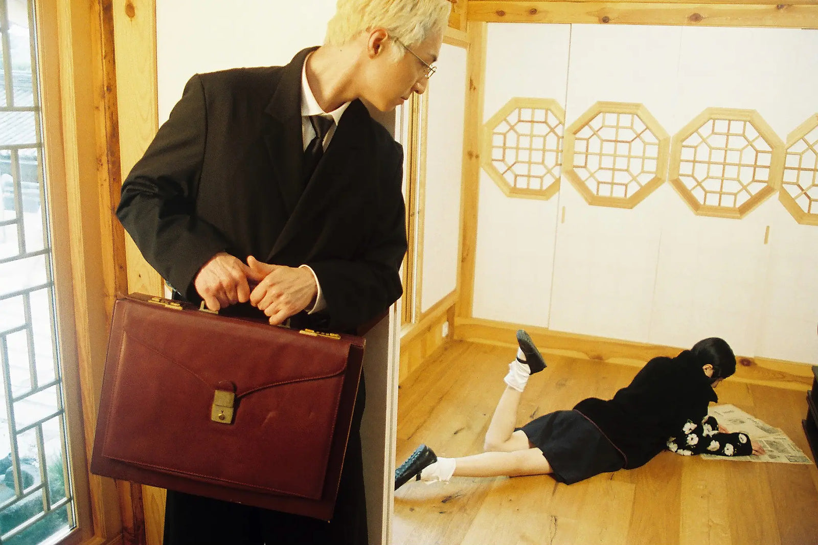 A man in a black suit holding a brown leather briefcase stands in a traditional Korean room, looking down at a person in Sundae School Mugunghwa Fleece lying on the wooden floor.