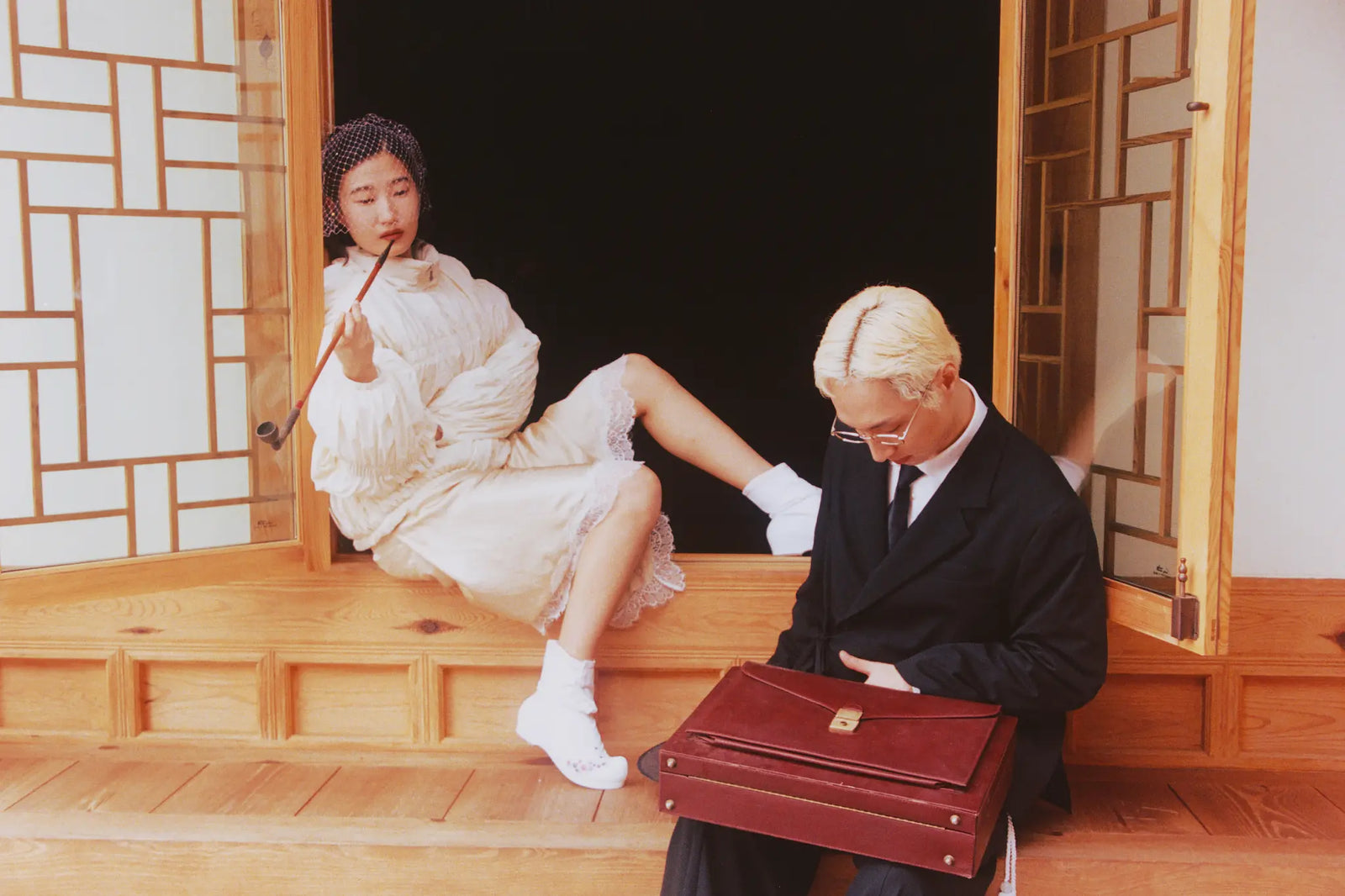 A man in a black suit and tie sits holding a dark red leather briefcase, while a woman in Sundae School Pleated Duvet Puffer lounges in a wooden doorway holding a pipe.