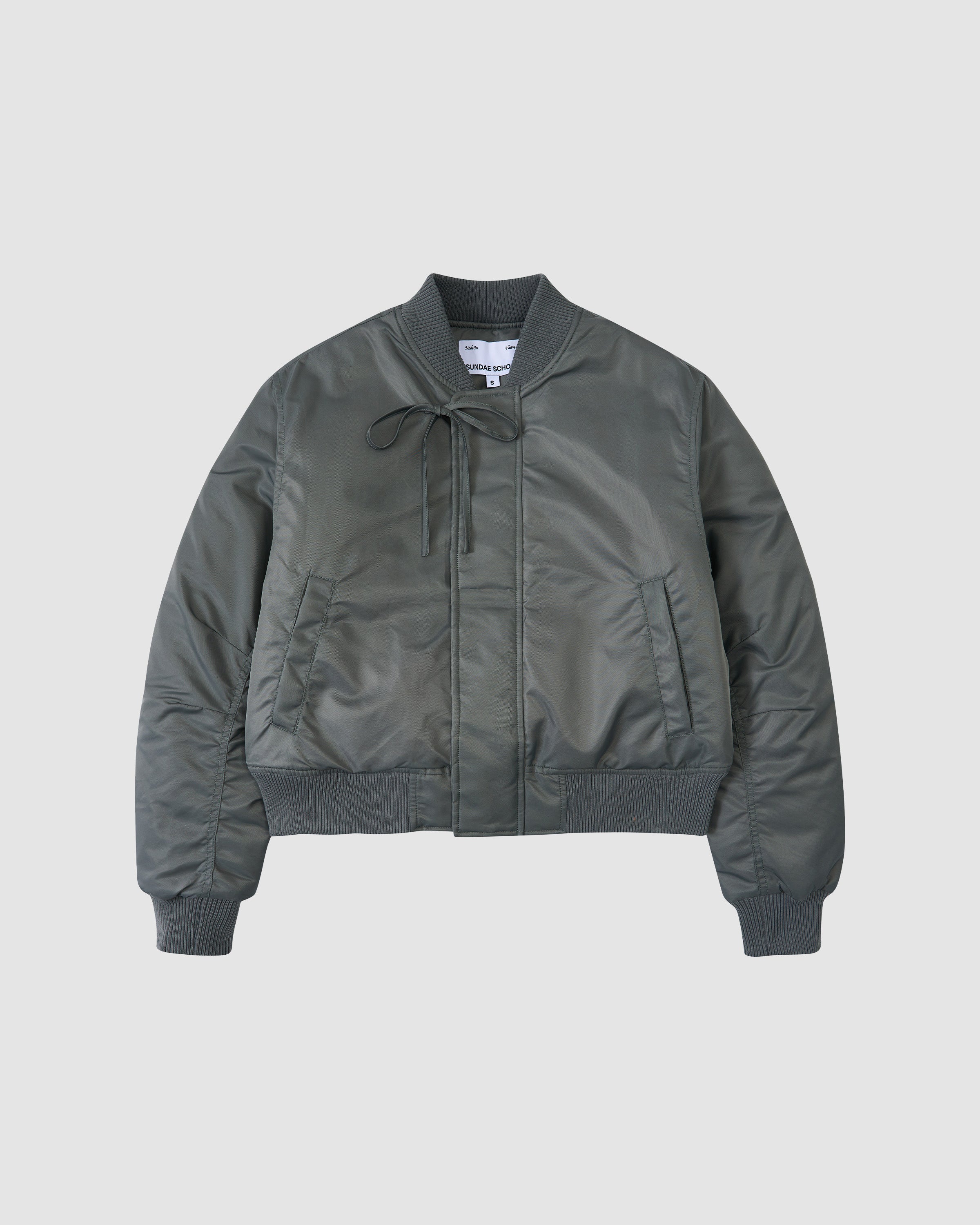The KHAKI FLOWER CHANNELED BOMBER by Sundae School is a dark green jacket with ribbed cuffs, collar, and hem, front zipper, side pockets, and a drawstring at the collar, shown against a plain light background.