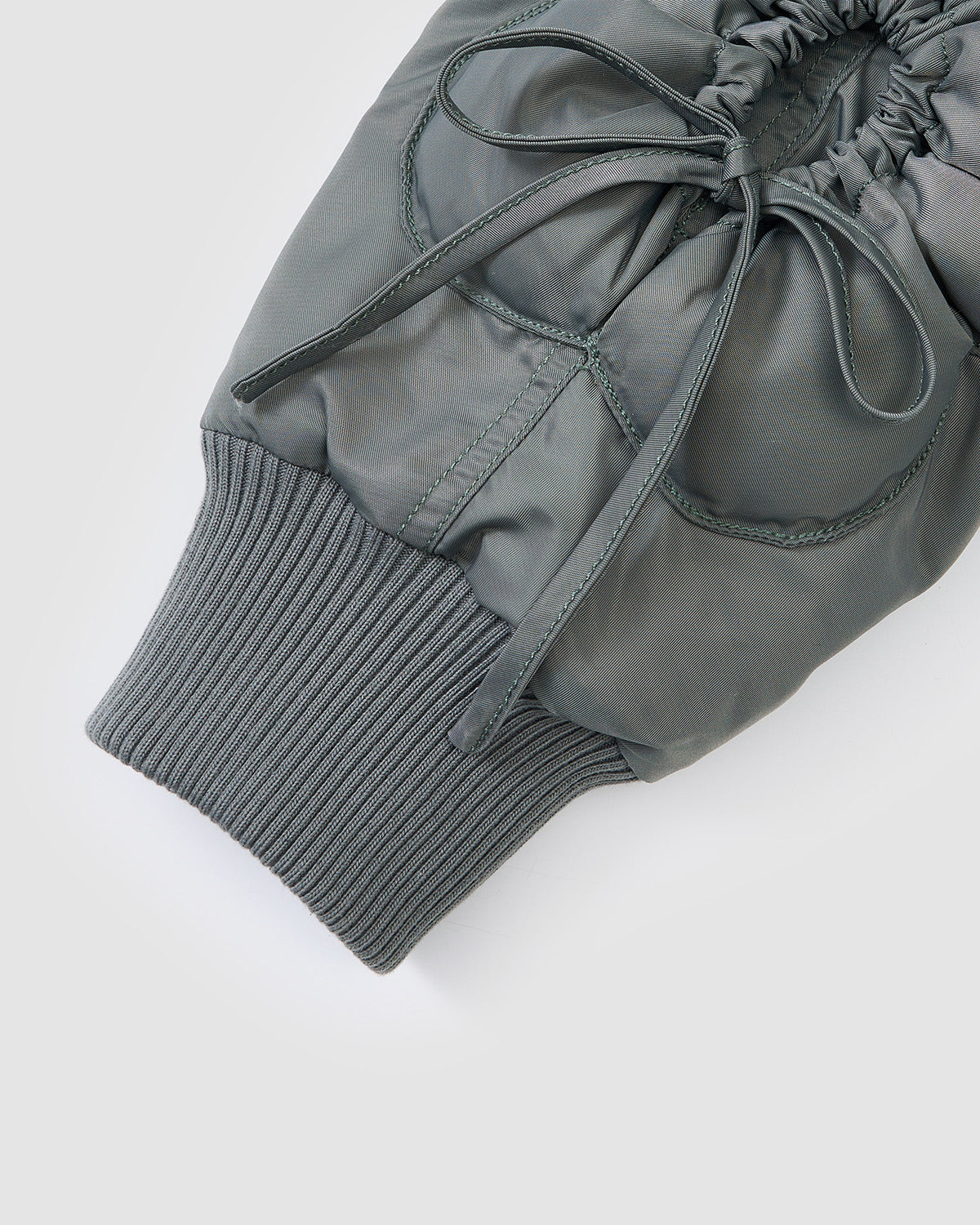 A close-up of the KHAKI FLOWER CHANNELED BOMBER by Sundae School shows the gray padded sleeve with a ribbed knit cuff and elastic drawstring detail near the wrist against a plain, light-colored background.