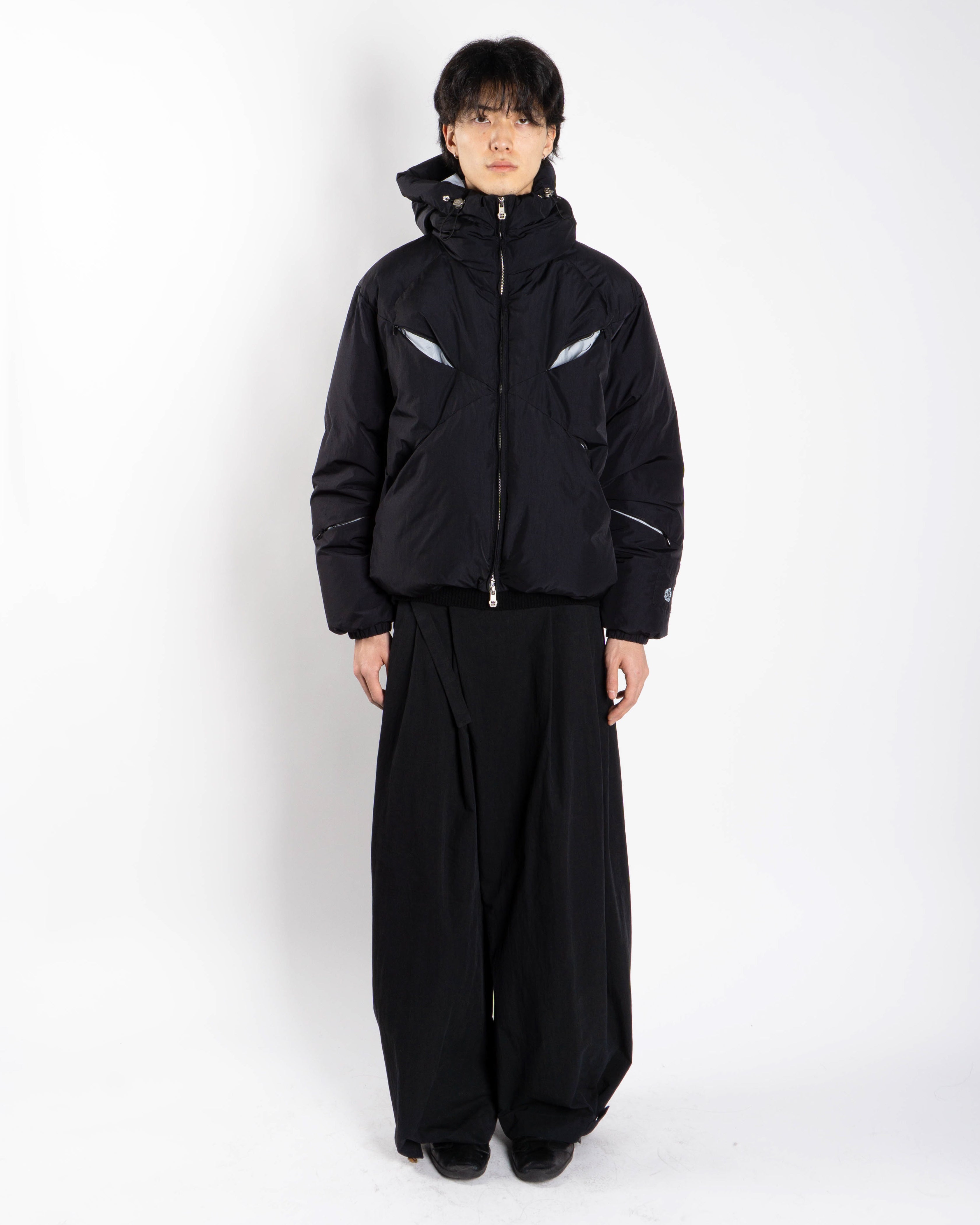 A person stands against a white background wearing the Sundae School BLACK SKY BLUE ORIGAMI PUFFER and wide-legged black pants, facing forward with hands at their sides.