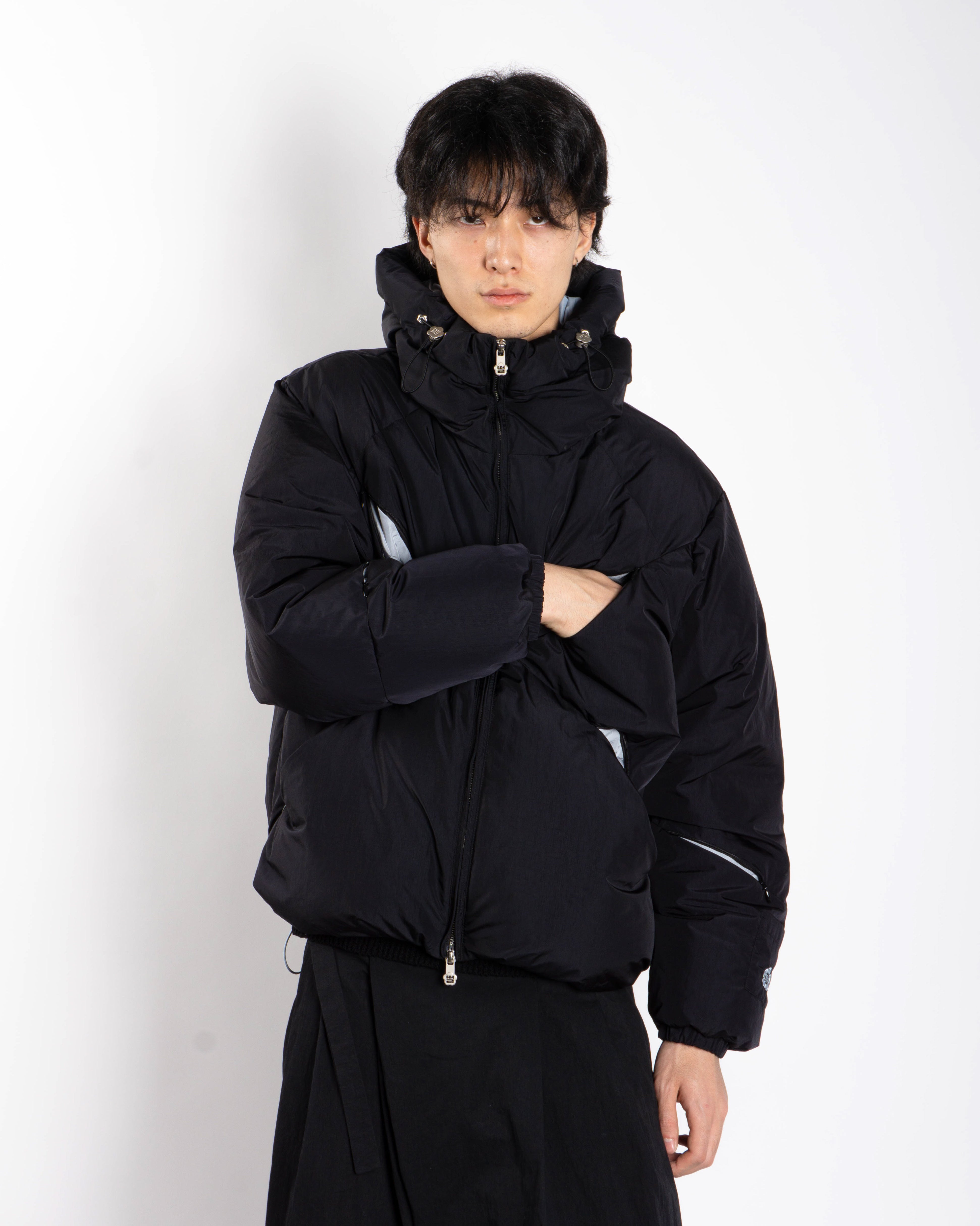 A person with short dark hair models the Sundae School BLACK SKY BLUE ORIGAMI PUFFER, featuring a high collar and hood, standing against a plain white background with one arm crossed over their chest.