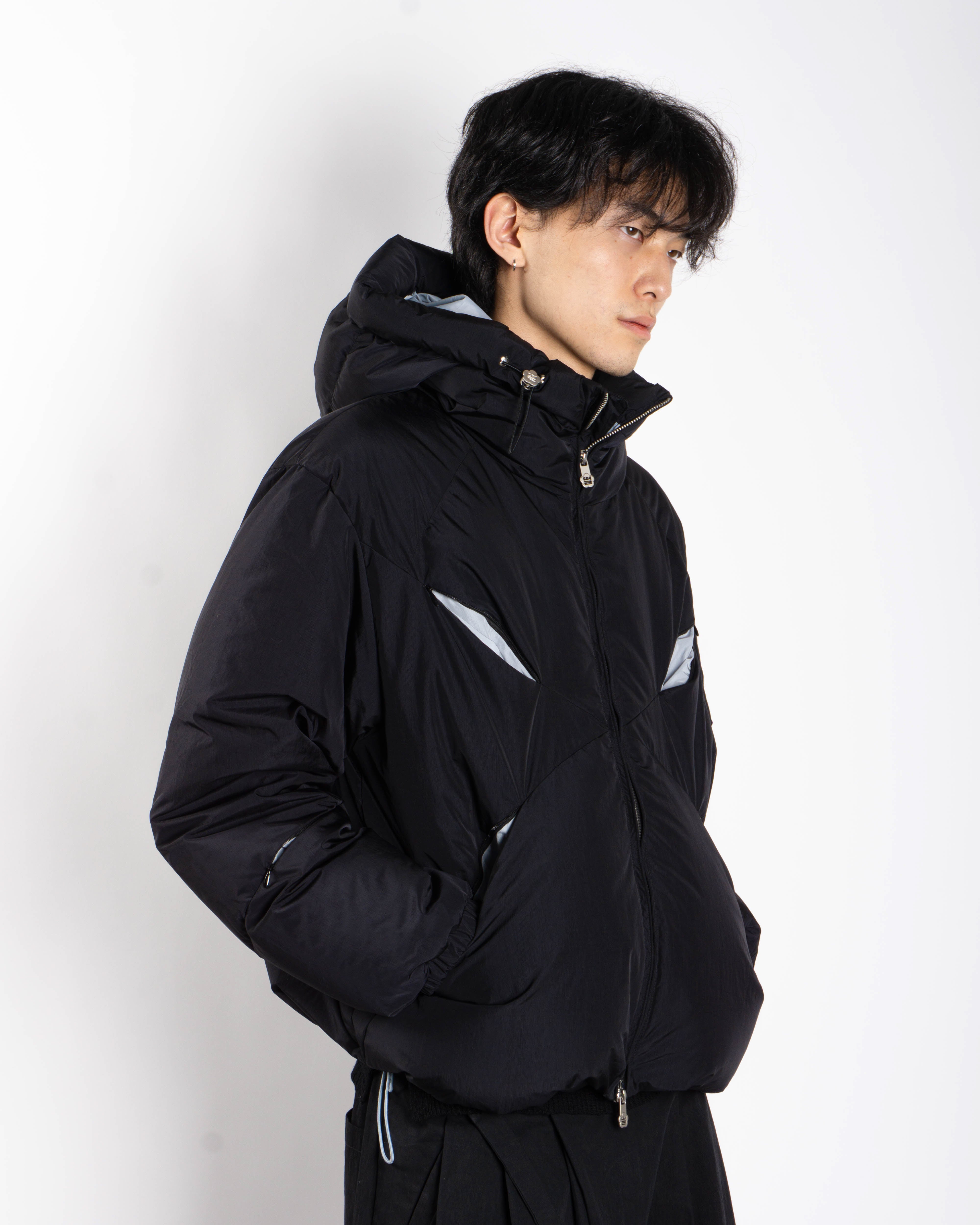 A person with dark hair wears the Sundae School BLACK SKY BLUE ORIGAMI PUFFER, featuring silver accents and a black hood, standing against a white background with hands in pockets, looking to the side.