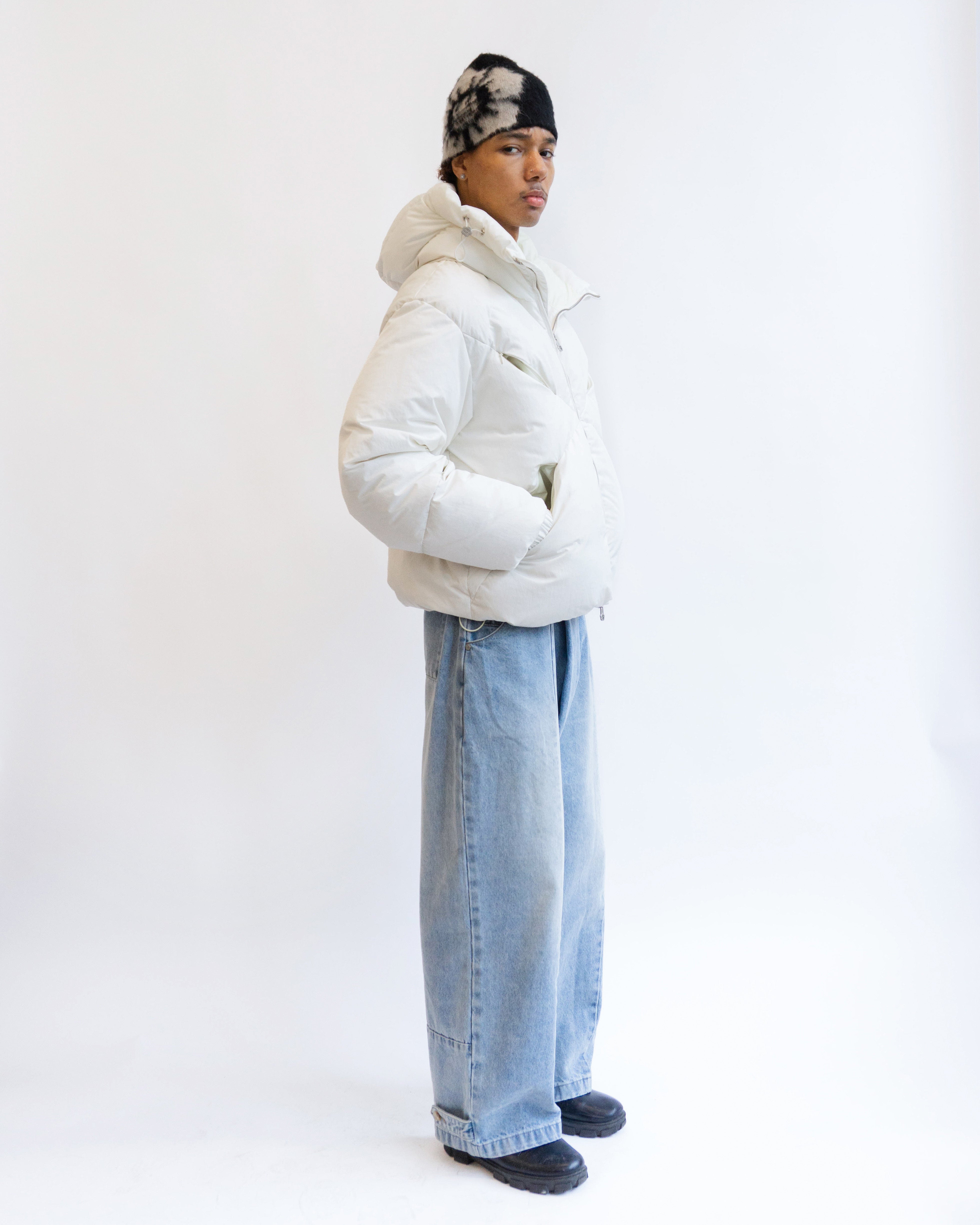 A person stands against a plain white background wearing the Sundae School IVORY LIME ORIGAMI PUFFER, wide-leg blue jeans, black sneakers, and a black and white beanie. Hands are in jacket pockets, gaze directed off-camera.