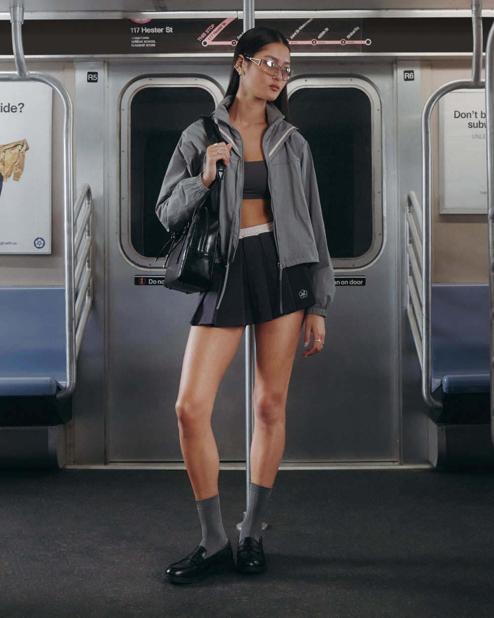A woman stands in a subway car, holding a black bag. She is wearing the GREY SAILOR WINDBREAKER by Sundae School, paired with a black sports bra, black shorts, and gray socks with loafers. The subway seats are empty and there are ads on the walls.