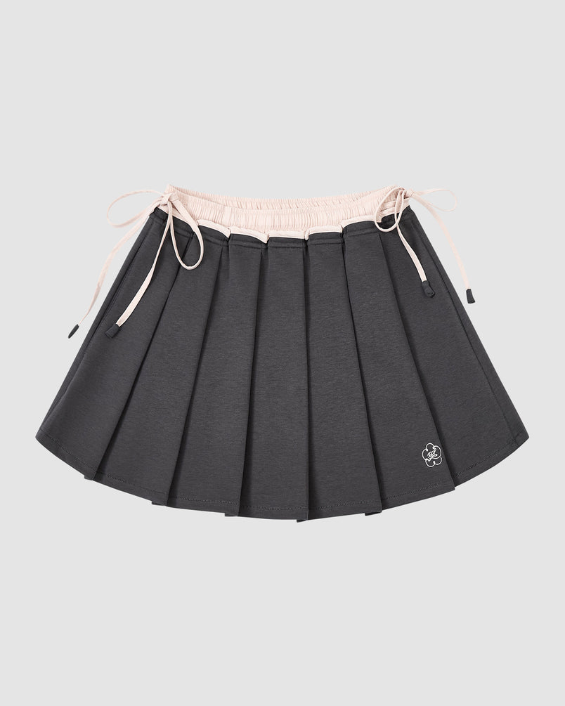 The CHARCOAL PLEATED JERSEY MINI SKIRT by Sundae School features dark gray knife pleats, a light pink elastic waistband with drawstrings, and a small white embroidered flower at the hem, shown against a plain light gray background.
